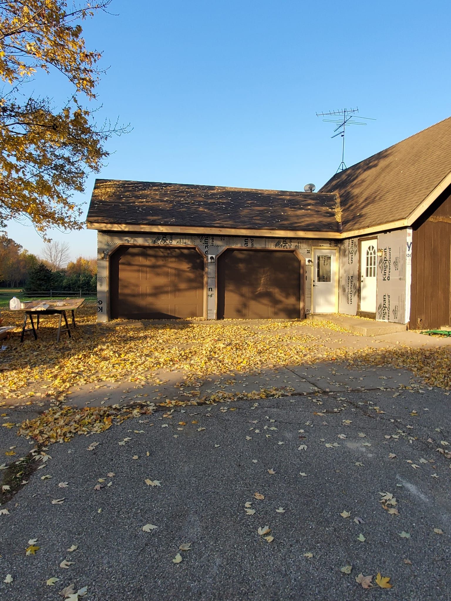 Autumn yard with a brown garage and white shed, covered in fallen leaves under a clear blue sky