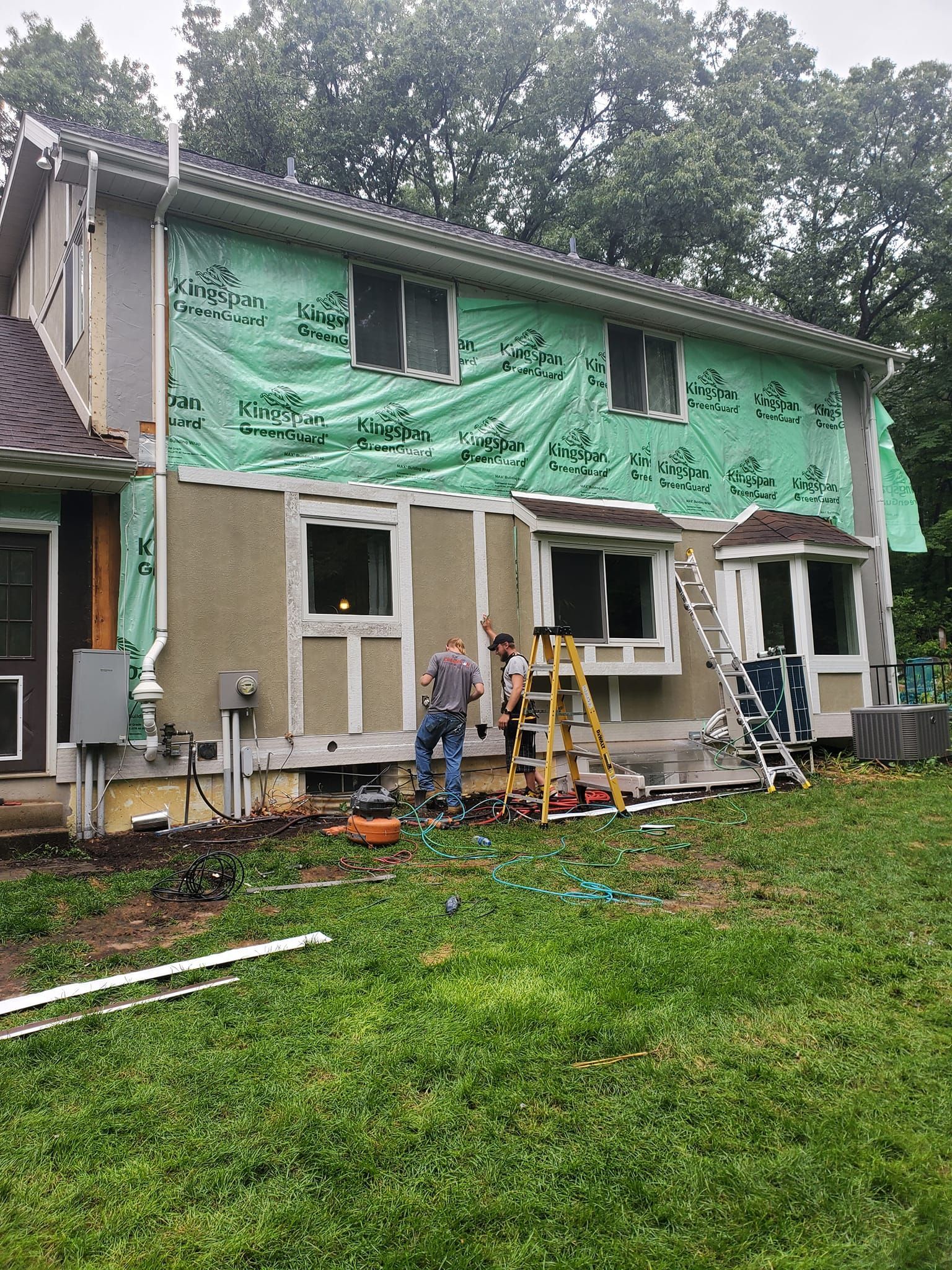 Two-story house under repair with green tarp on upper siding and workers on ladders in front yard