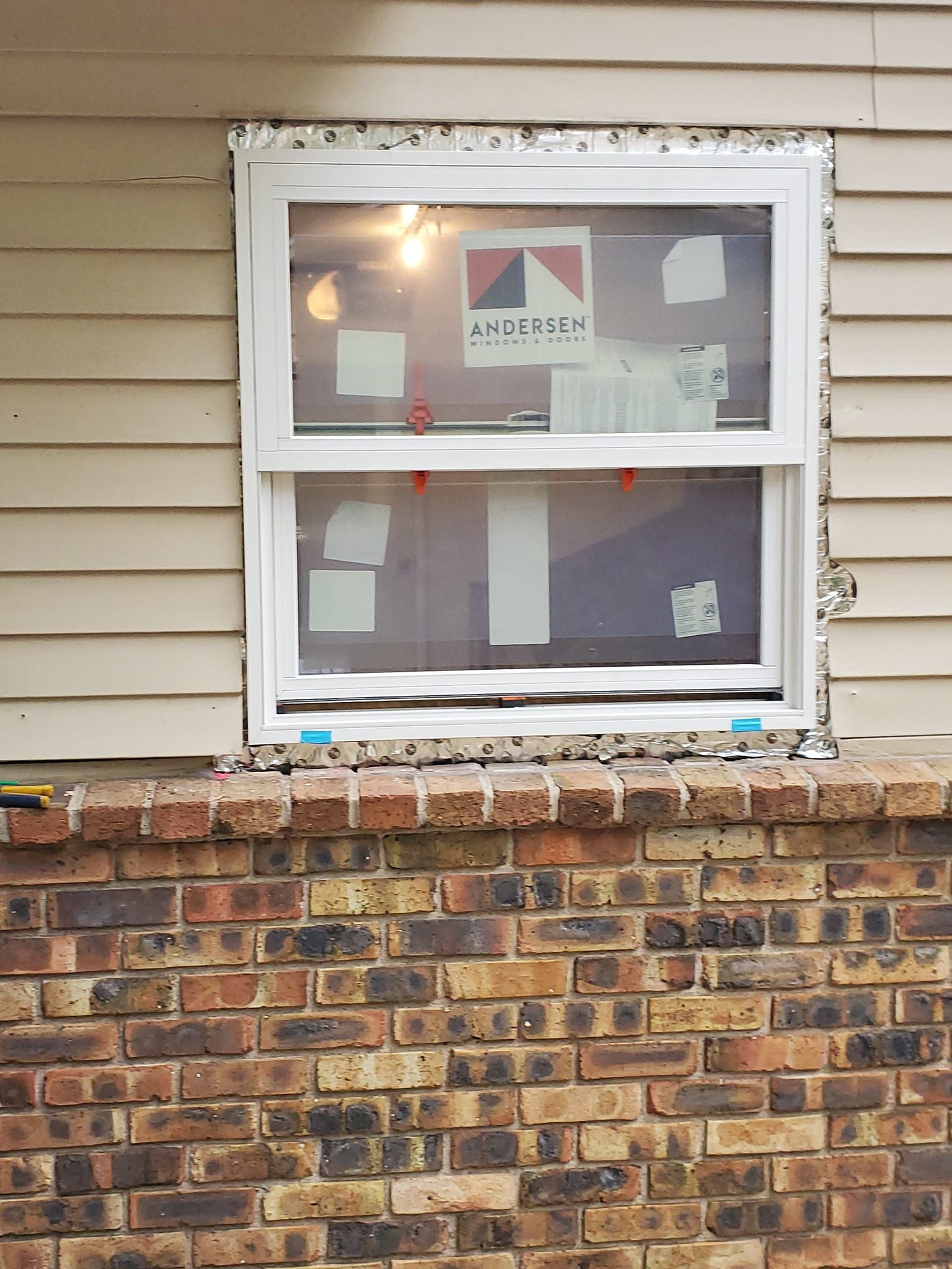 House window with a white frame and a sign inside, set above a brick wall and tan siding.
