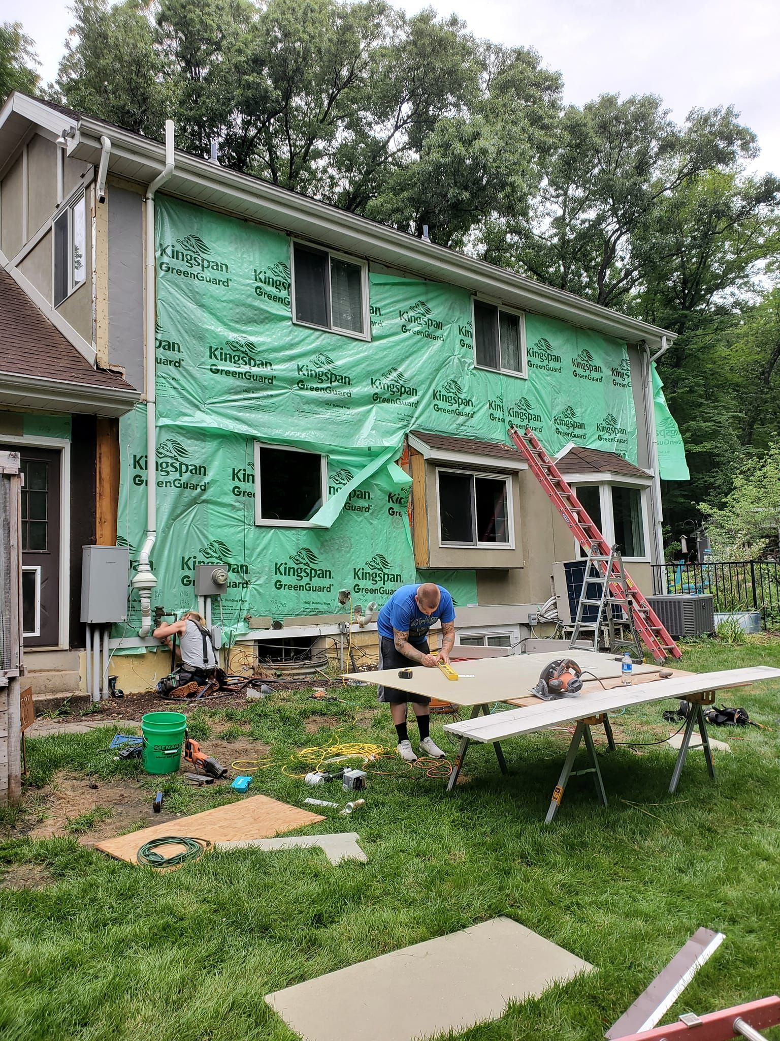 Workers on a two-story house with green wrap and ladders during exterior renovation