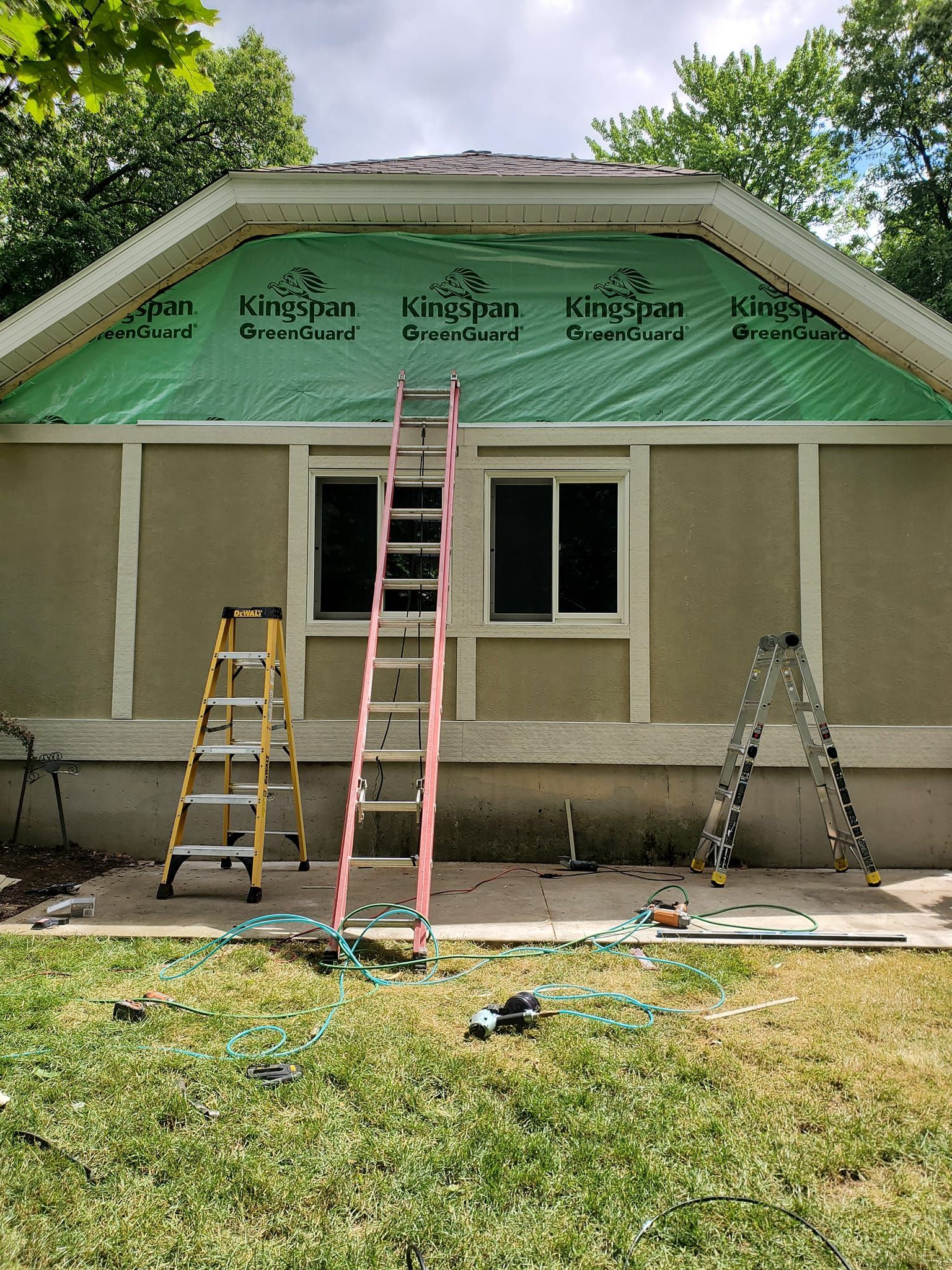 House exterior with green roof under construction, ladders on the front lawn, and exposed insulation under the eaves