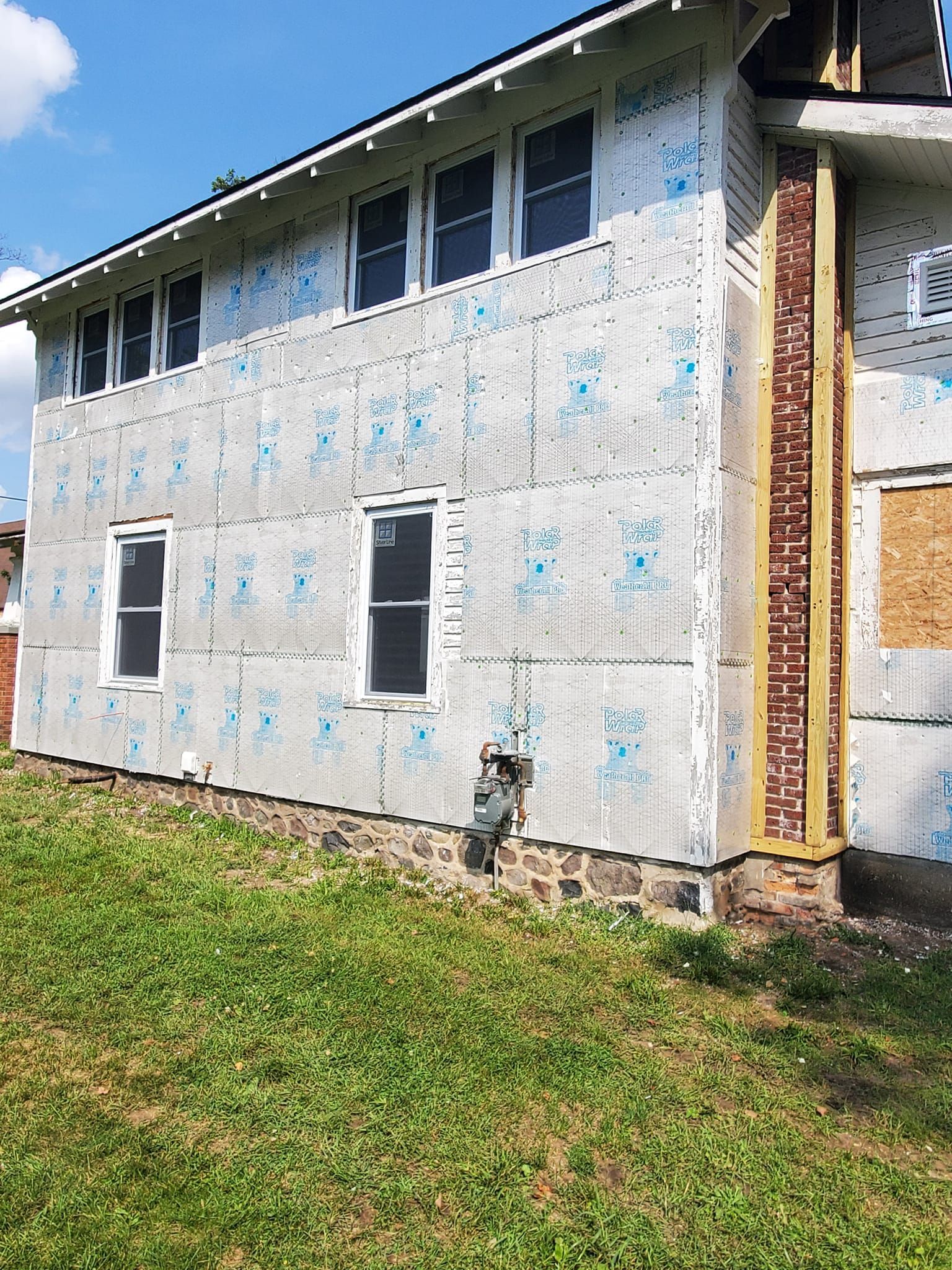 Two-story house under construction with white house wrap, blue sky, and grassy yard