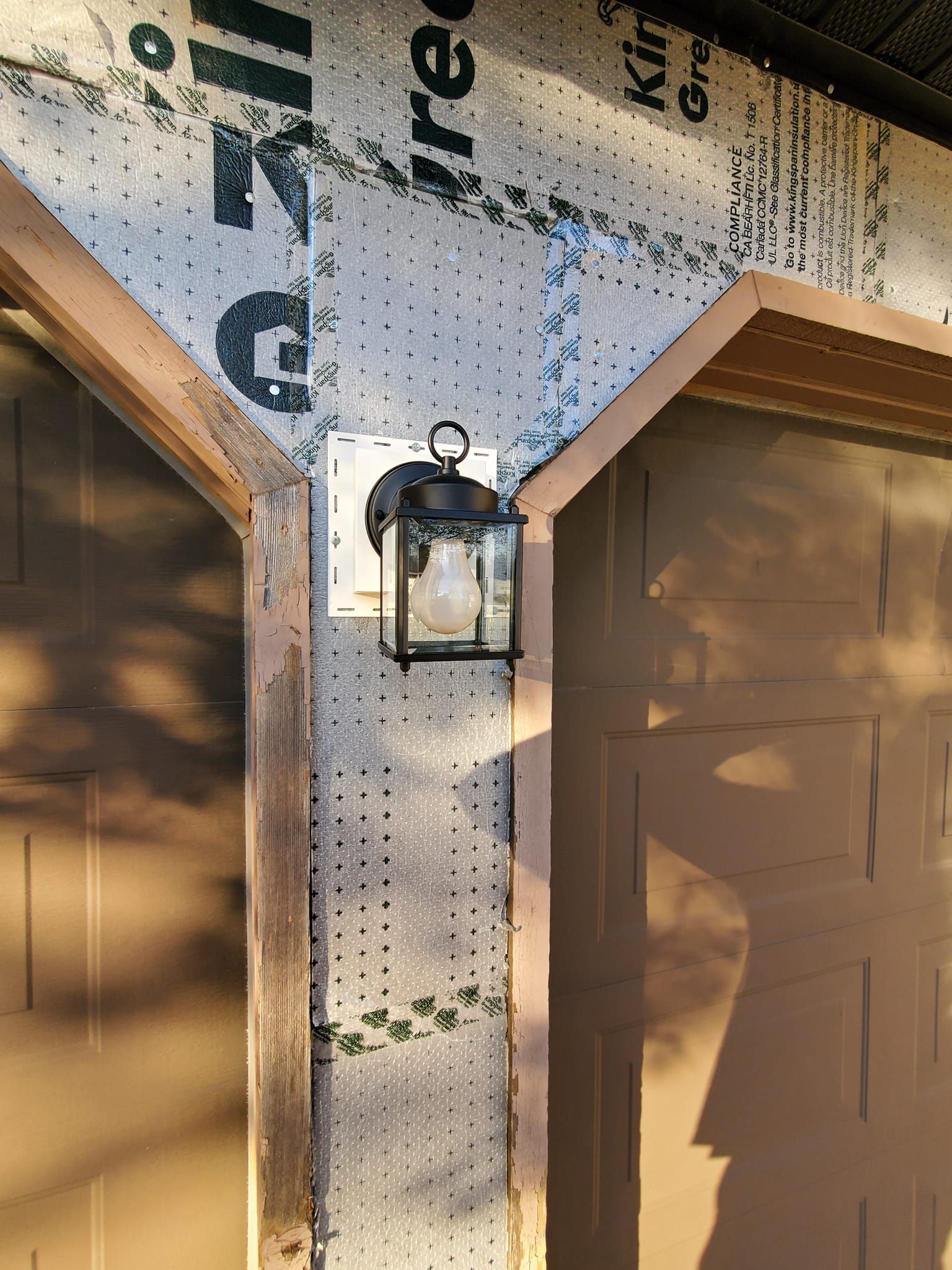 Outdoor wall lantern between tan garage doors under unfinished house wrap, with wood framing visible