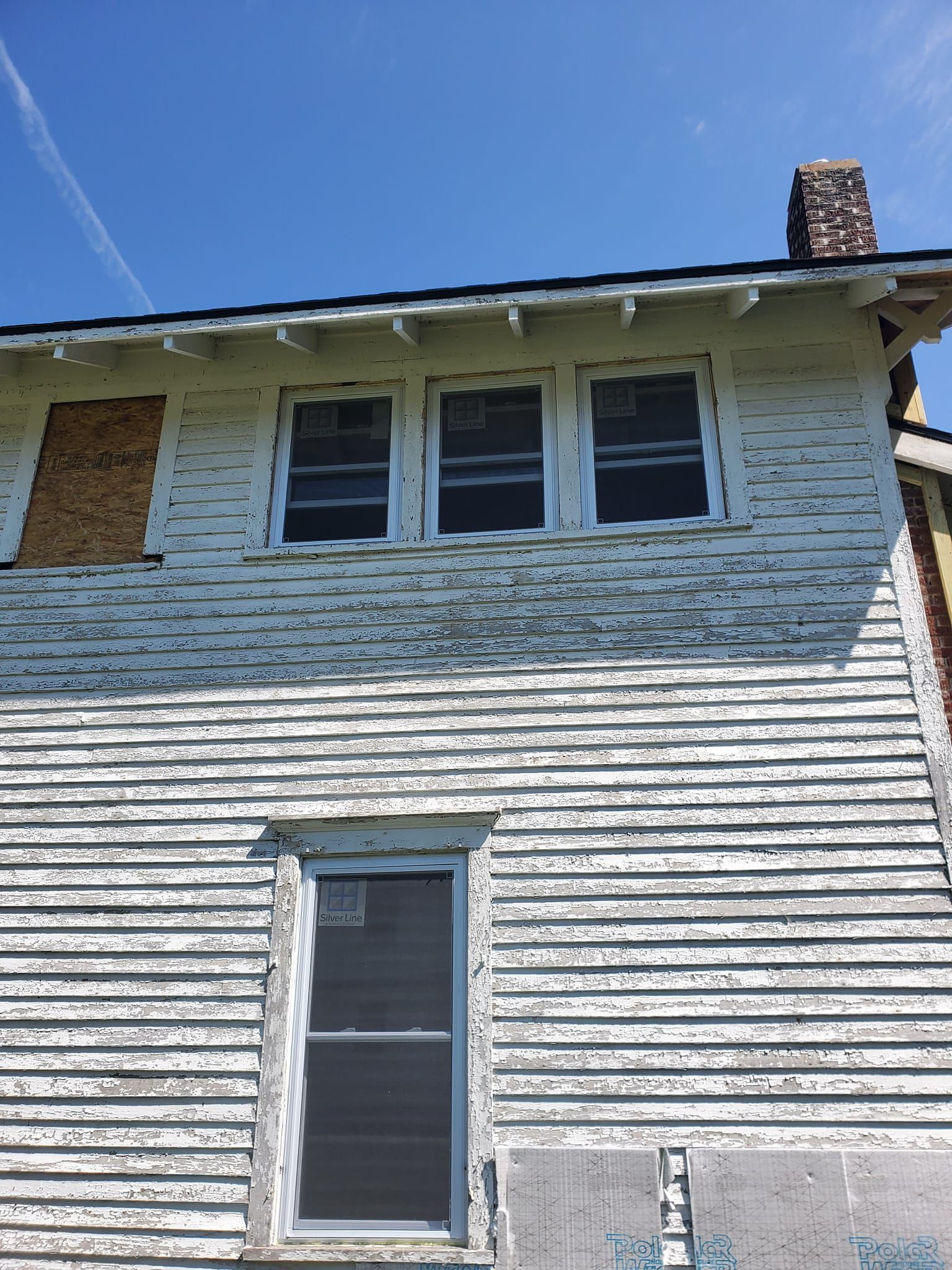 Weathered white house exterior with four upstairs windows, a narrow lower window, and a brick chimney under a blue sky