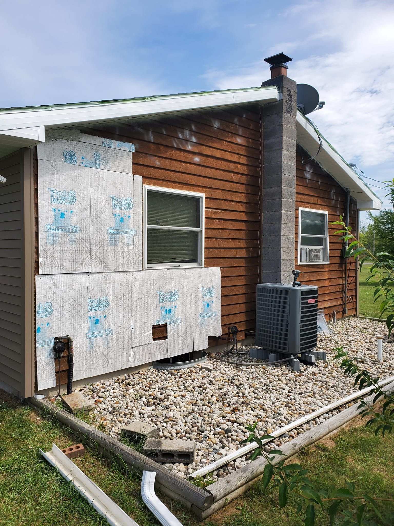 Damaged exterior wall with peeling panels, a small window, and utility equipment beside a gravel bed