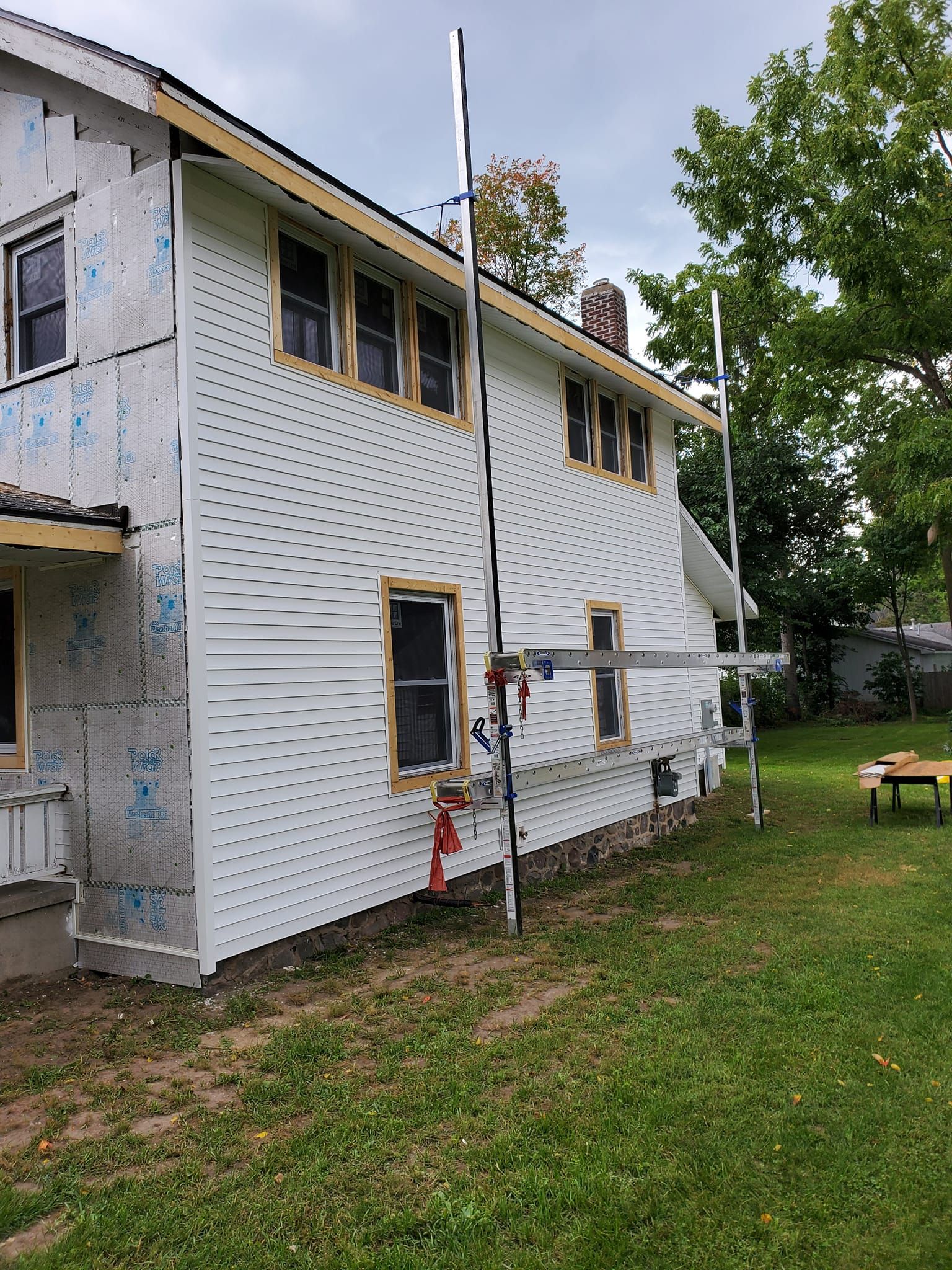 Two-story house under renovation with white siding, scaffolding, and a ladder beside the lawn