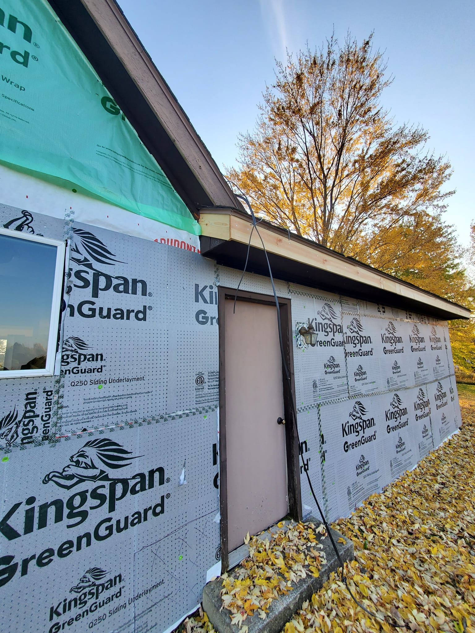 Partially built house exterior with white house wrap, brown door, and autumn tree beside it