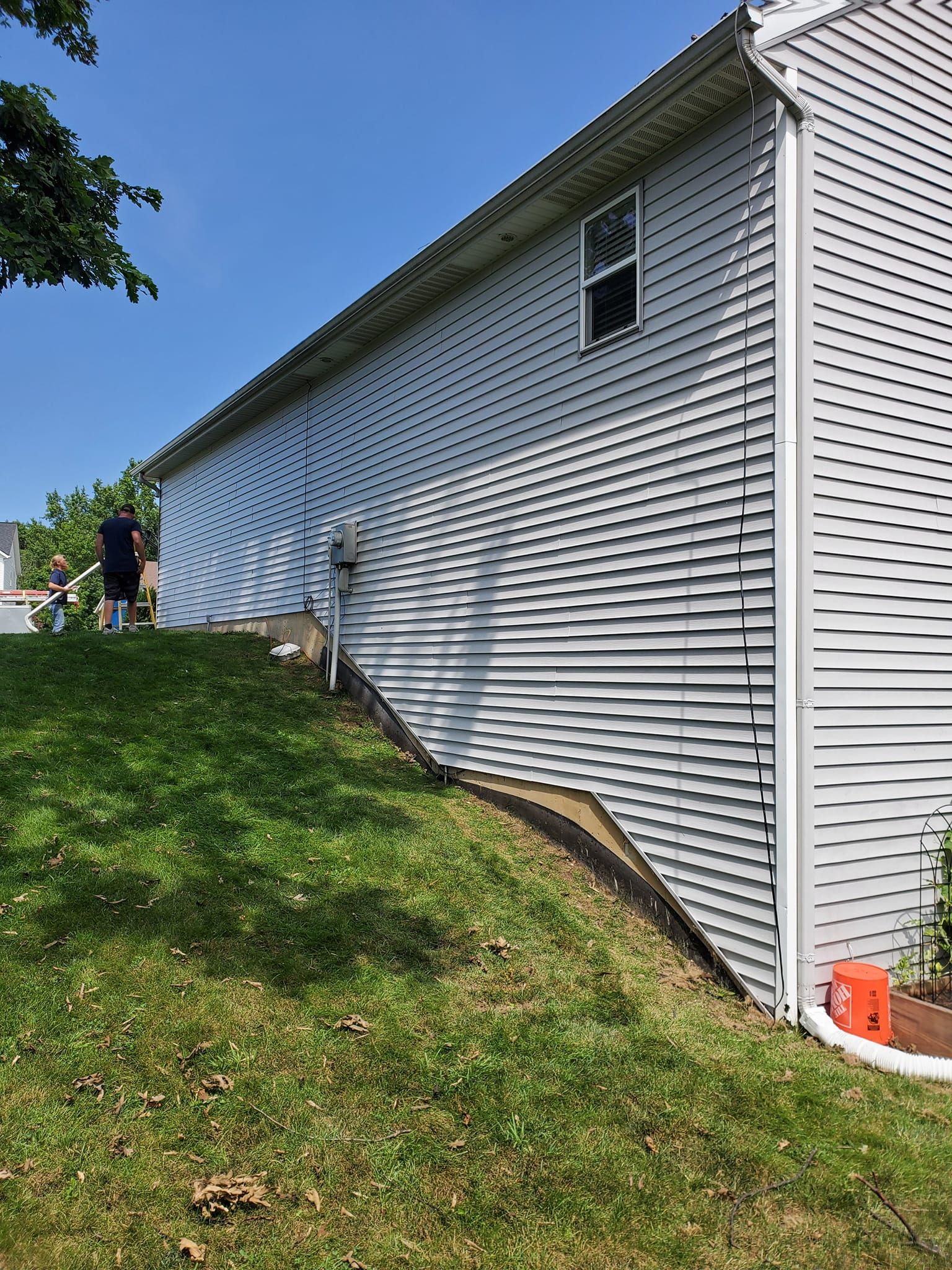 Side of white house with vinyl siding, grassy slope, and two people near the left edge under a blue sky.