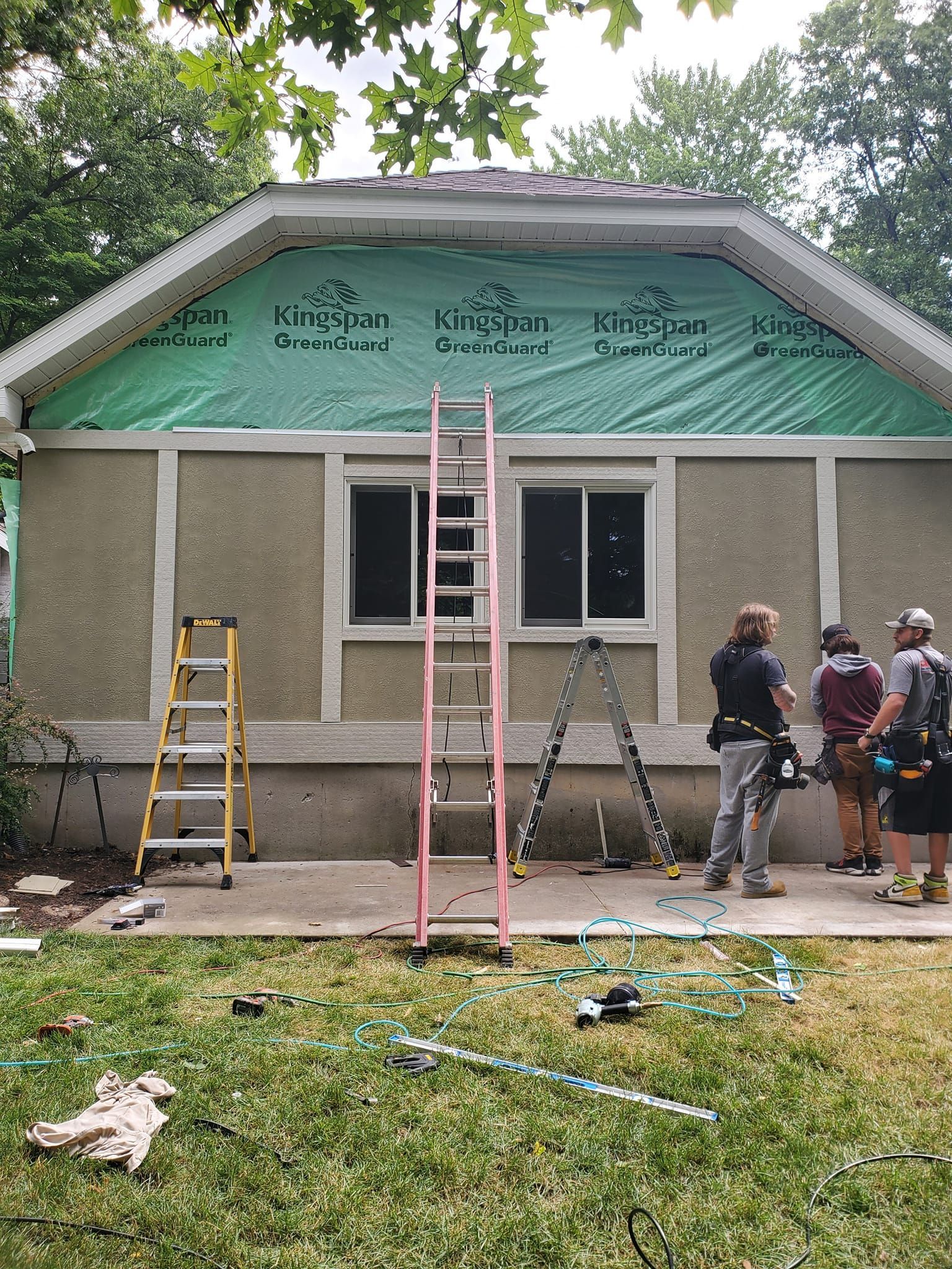 Workers renovating a house exterior, with ladders and green house wrap under the roofline.