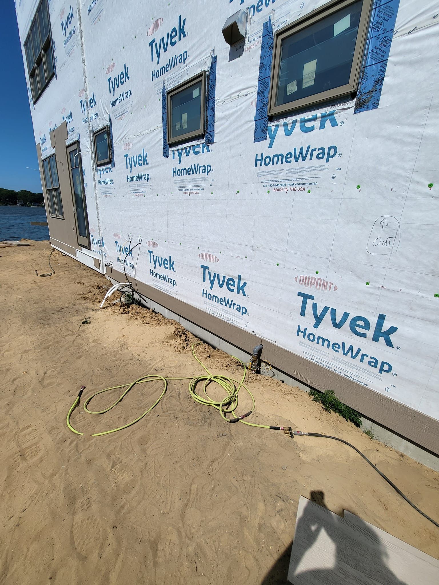 Construction site wall wrapped in Tyvek near a sandy shoreline, with windows installed and a worker’s shadow visible.