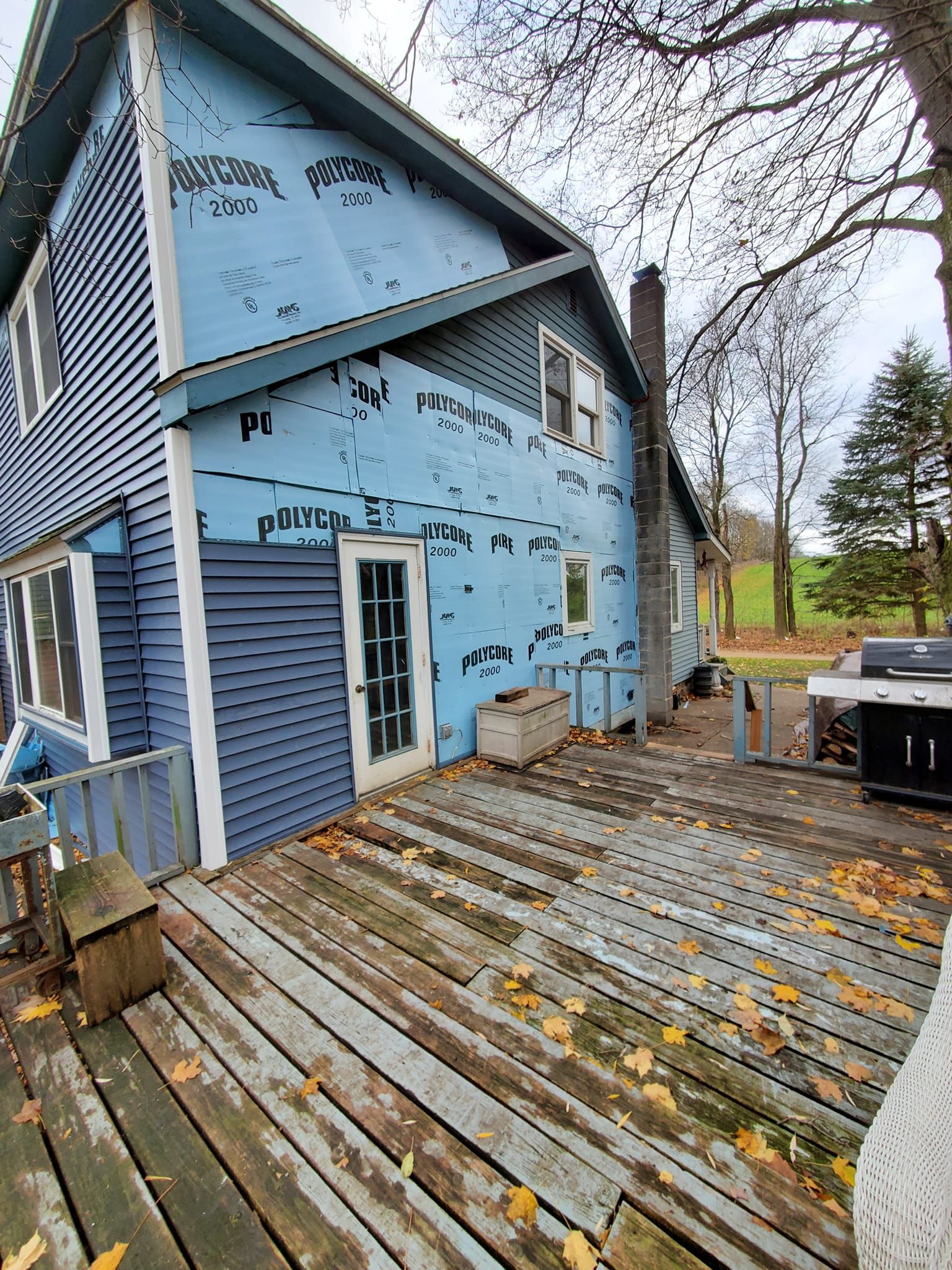 Blue house with exposed sheathing and a weathered wooden deck beside a yard with trees