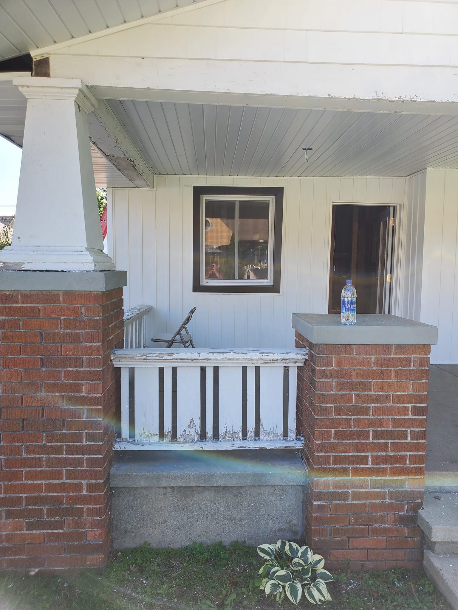 Front porch with brick columns, white siding, a window, railing, and a water bottle on the ledge.