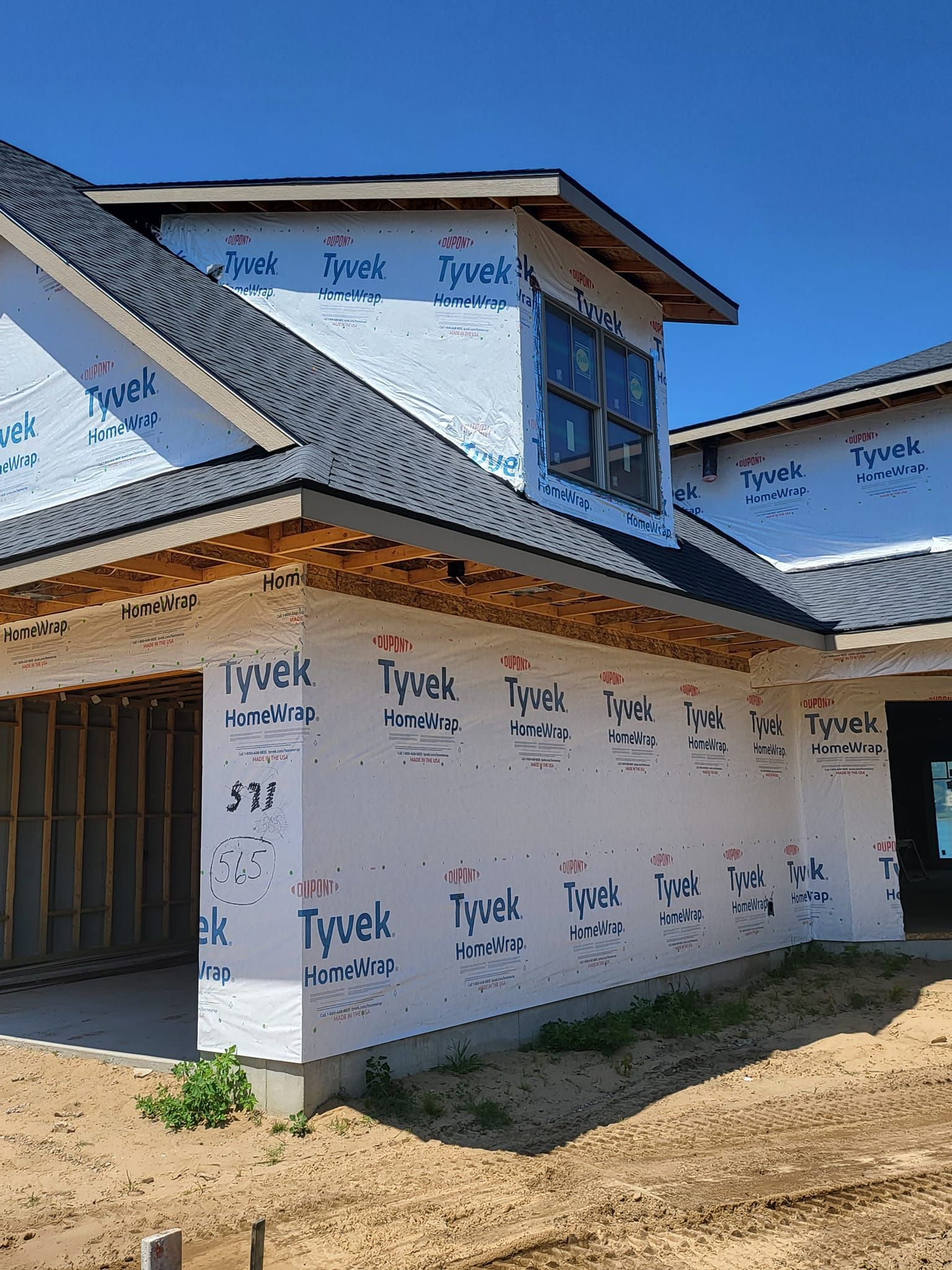 New house exterior under construction with Tyvek wrap, sloped roof, and unfinished dirt yard