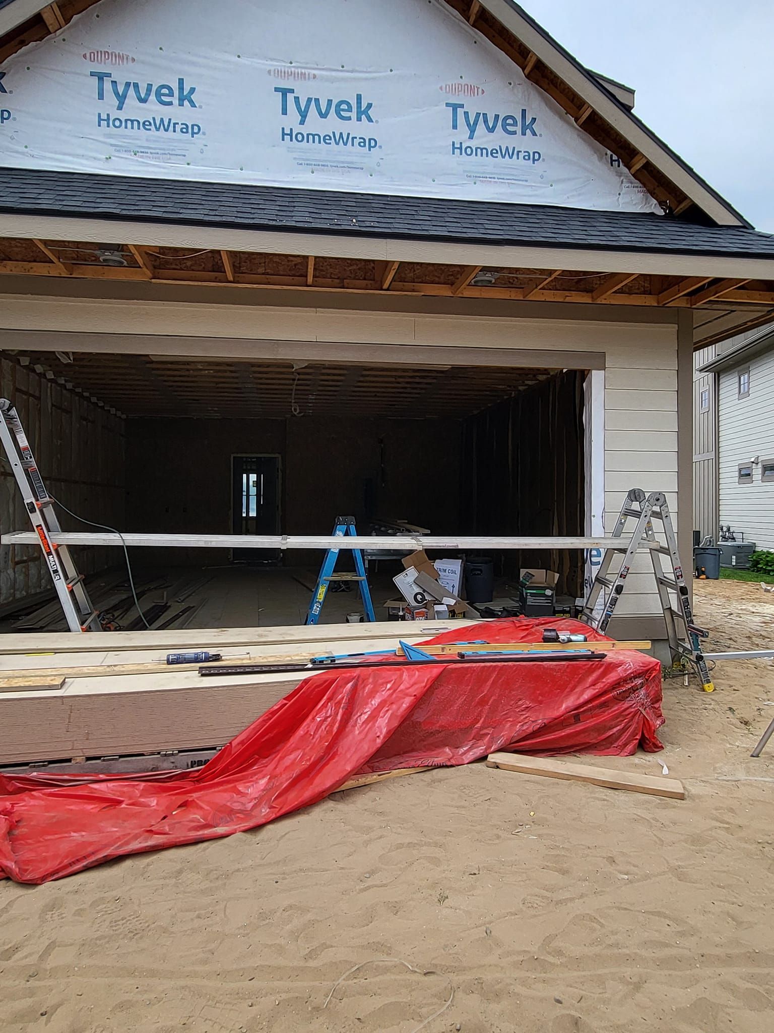 Garage under construction with Tyvek wrap, ladder, tools, and red tarp at the entrance