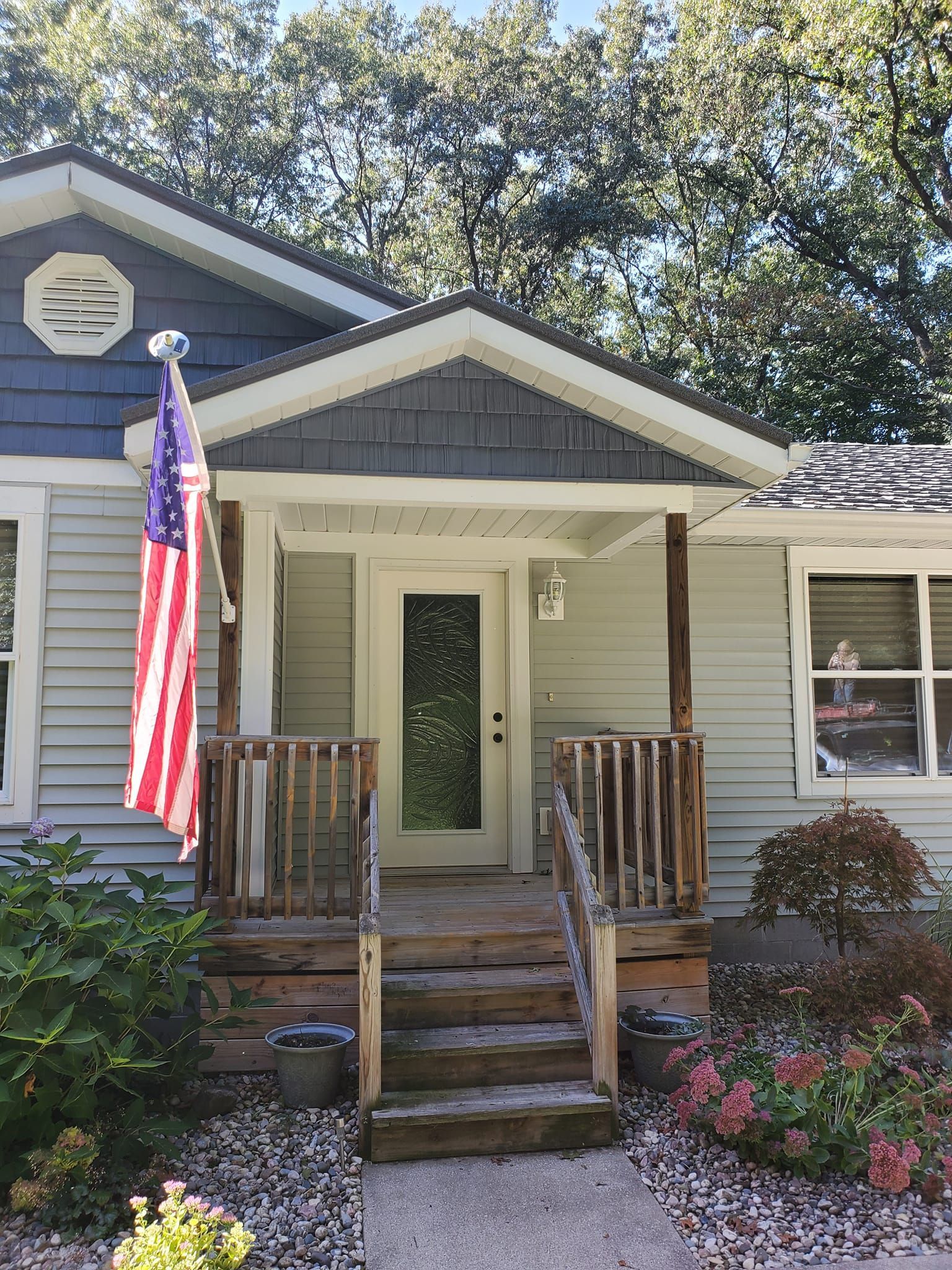 Front porch of a blue house with an American flag, steps, white trim, and flower beds.