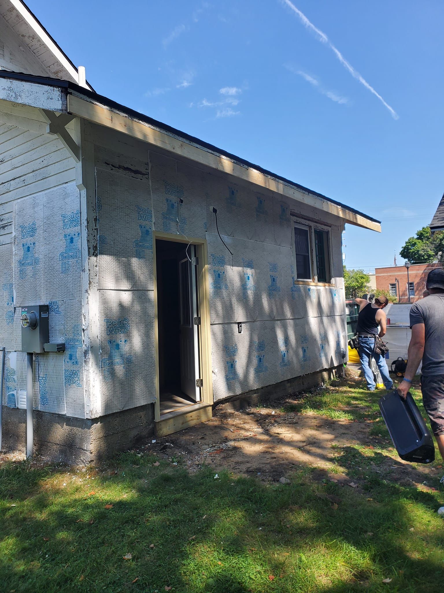 Small weathered building with open door, lawn in front, and people working outside under a blue sky.