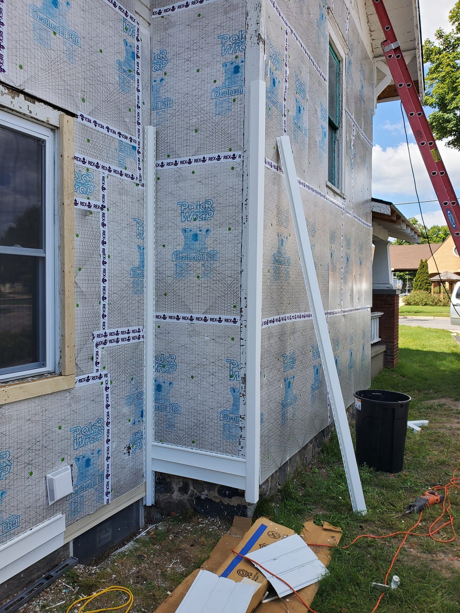 Partially wrapped house exterior with insulation panels, ladder, and construction materials on the ground.