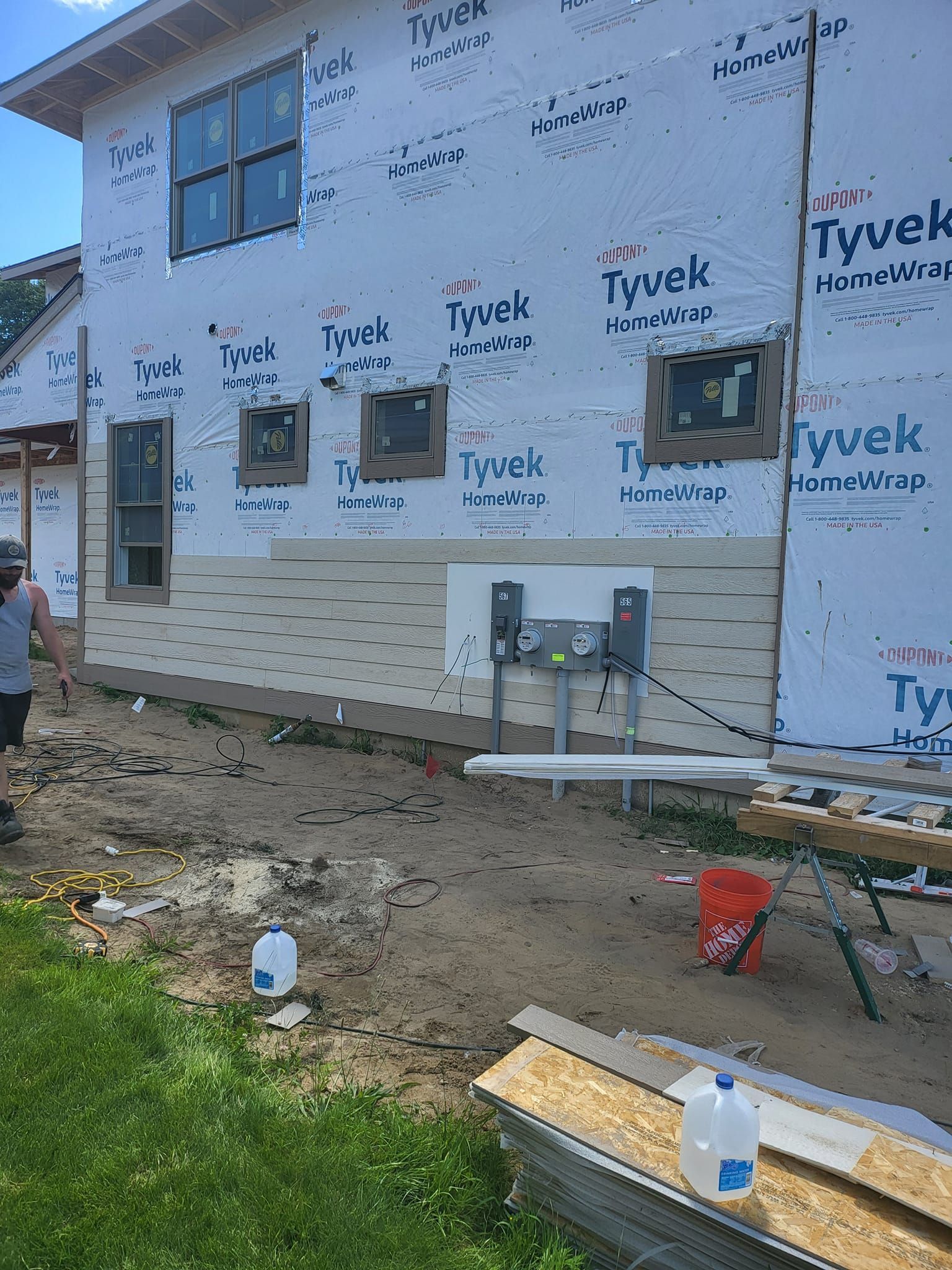 Partially built house wrapped in Tyvek, with exposed windows, utility boxes, and construction debris outside.