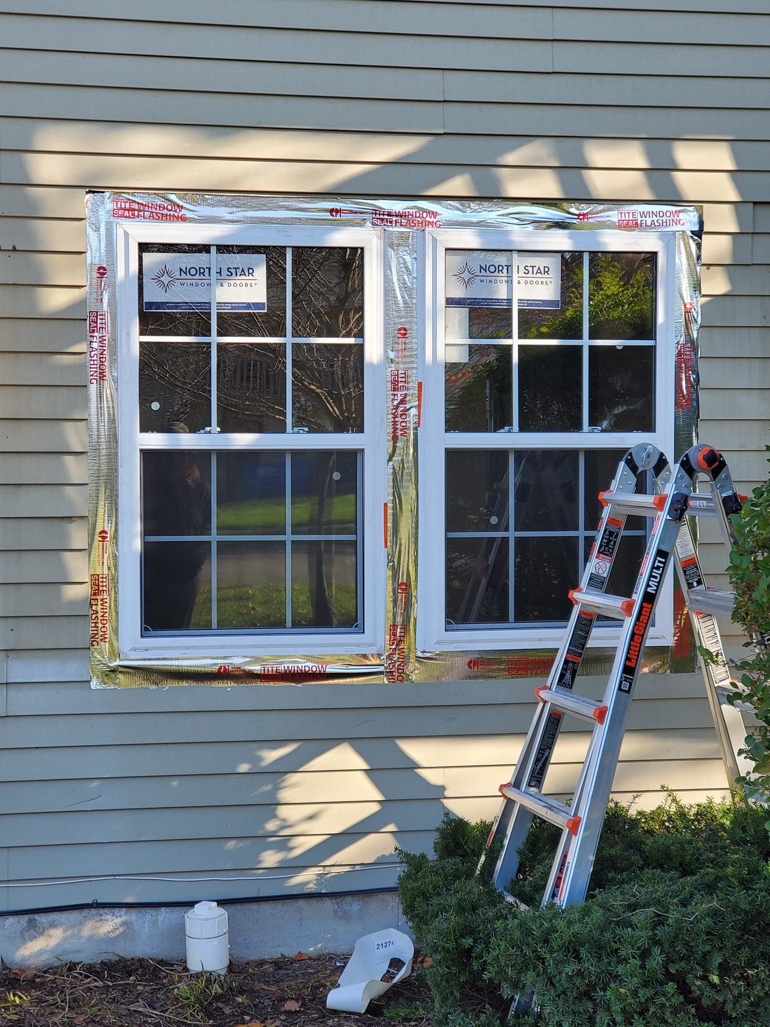 House window framed with white trim and festive garland, with a ladder leaning beside it on the exterior wall.