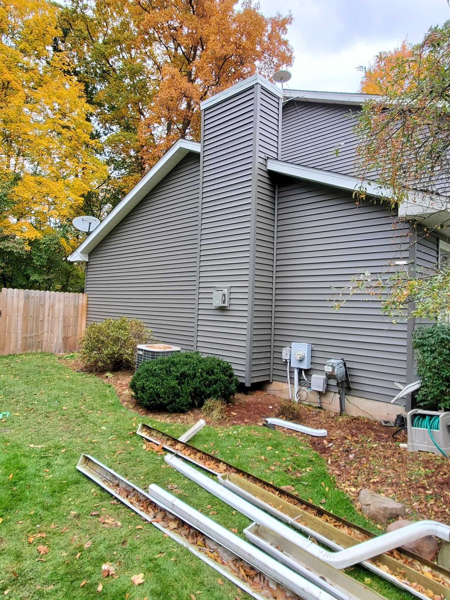 Damaged white siding and detached gutter pieces beside a house with autumn trees and a wooden fence