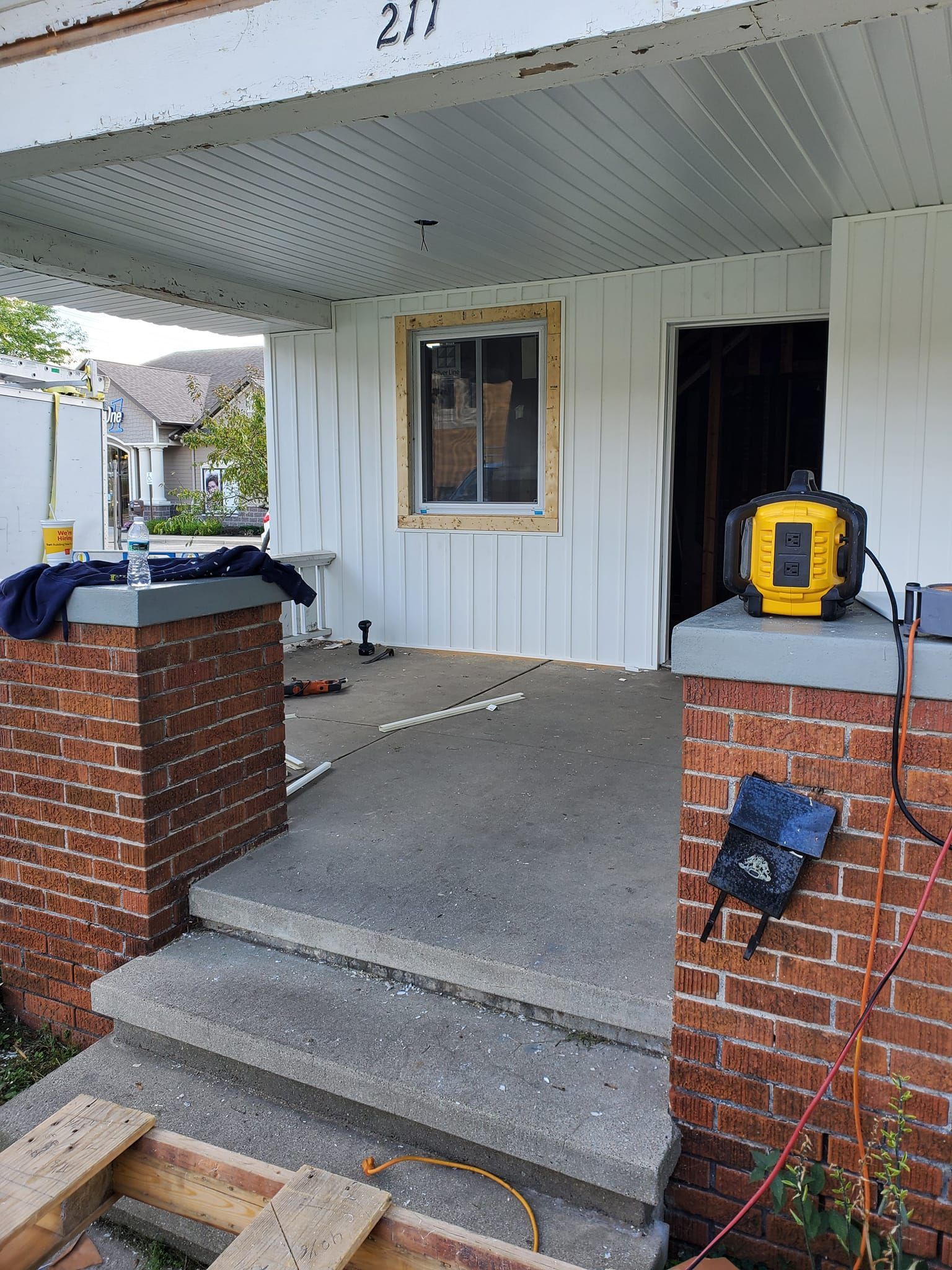 Front porch under renovation with concrete steps, brick pillars, white siding, and work tools on the ledge