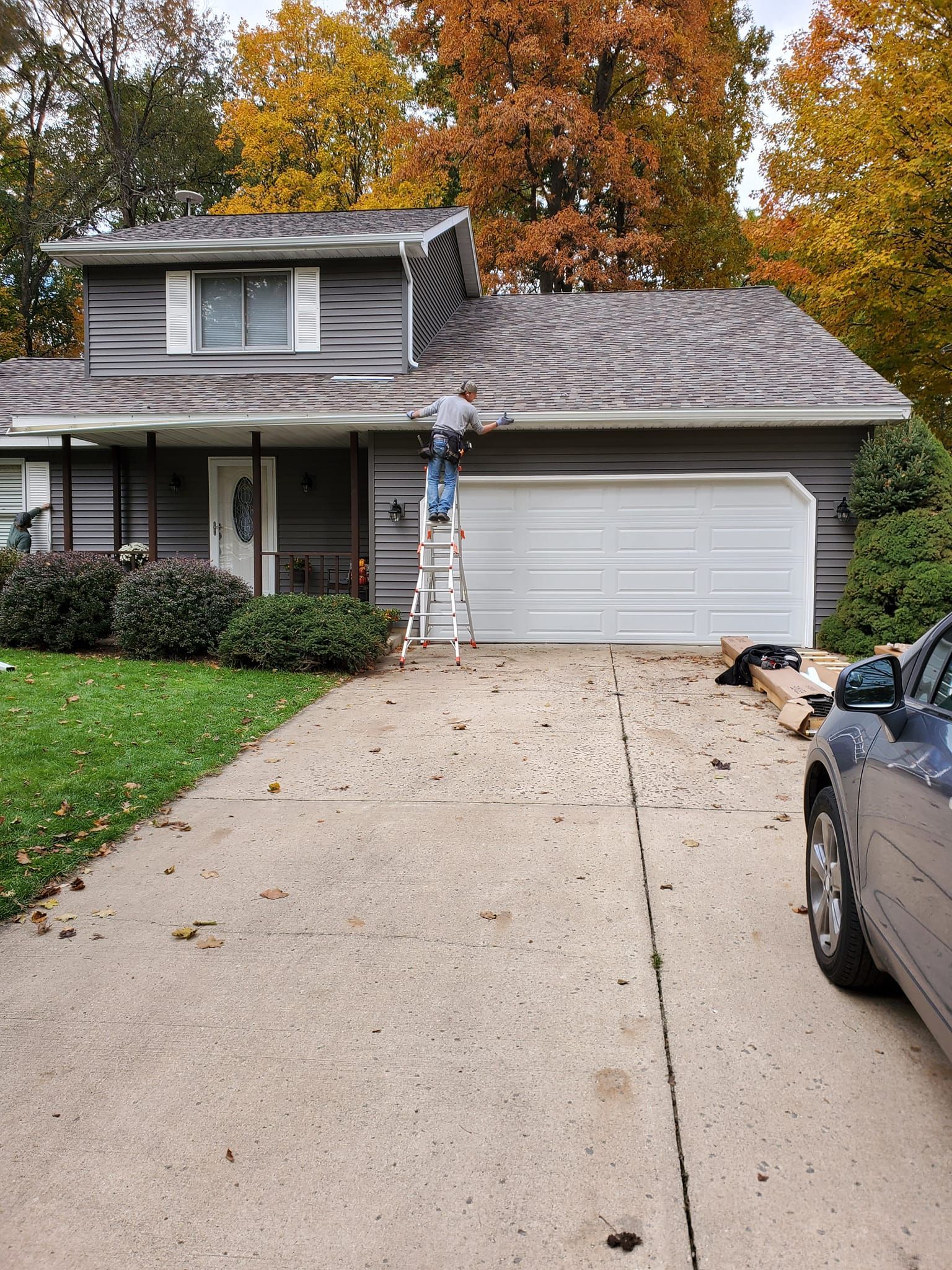 Suburban house with two-car garage, autumn trees, and a concrete driveway lined by parked cars