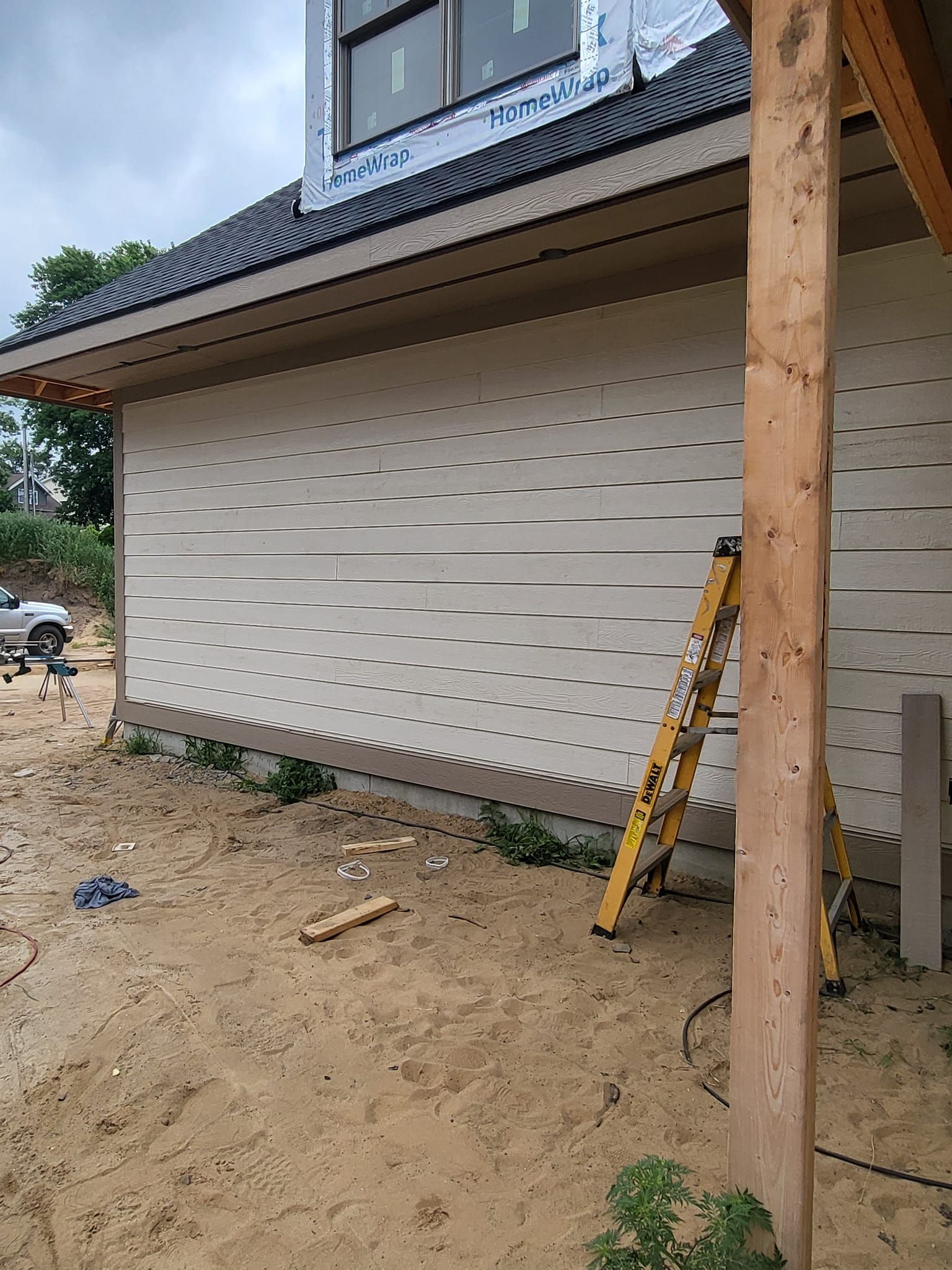 New house exterior with beige siding, sloped roof, wooden post, and a ladder on sandy ground.