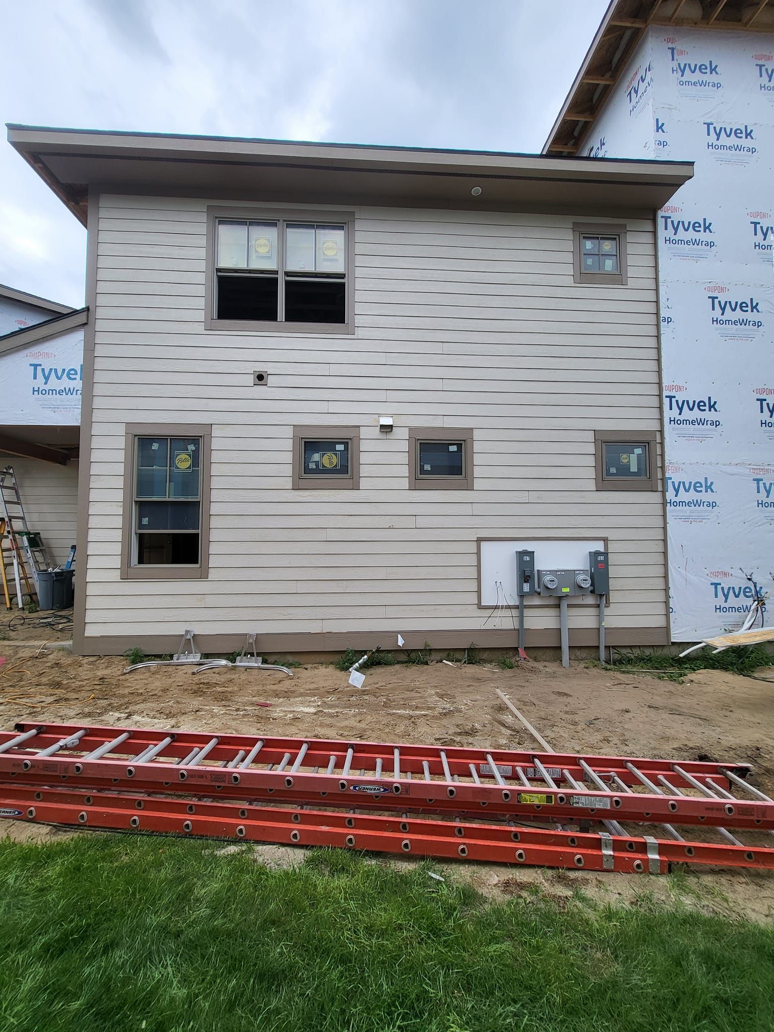 Two-story house exterior with beige siding, windows, and construction materials in front