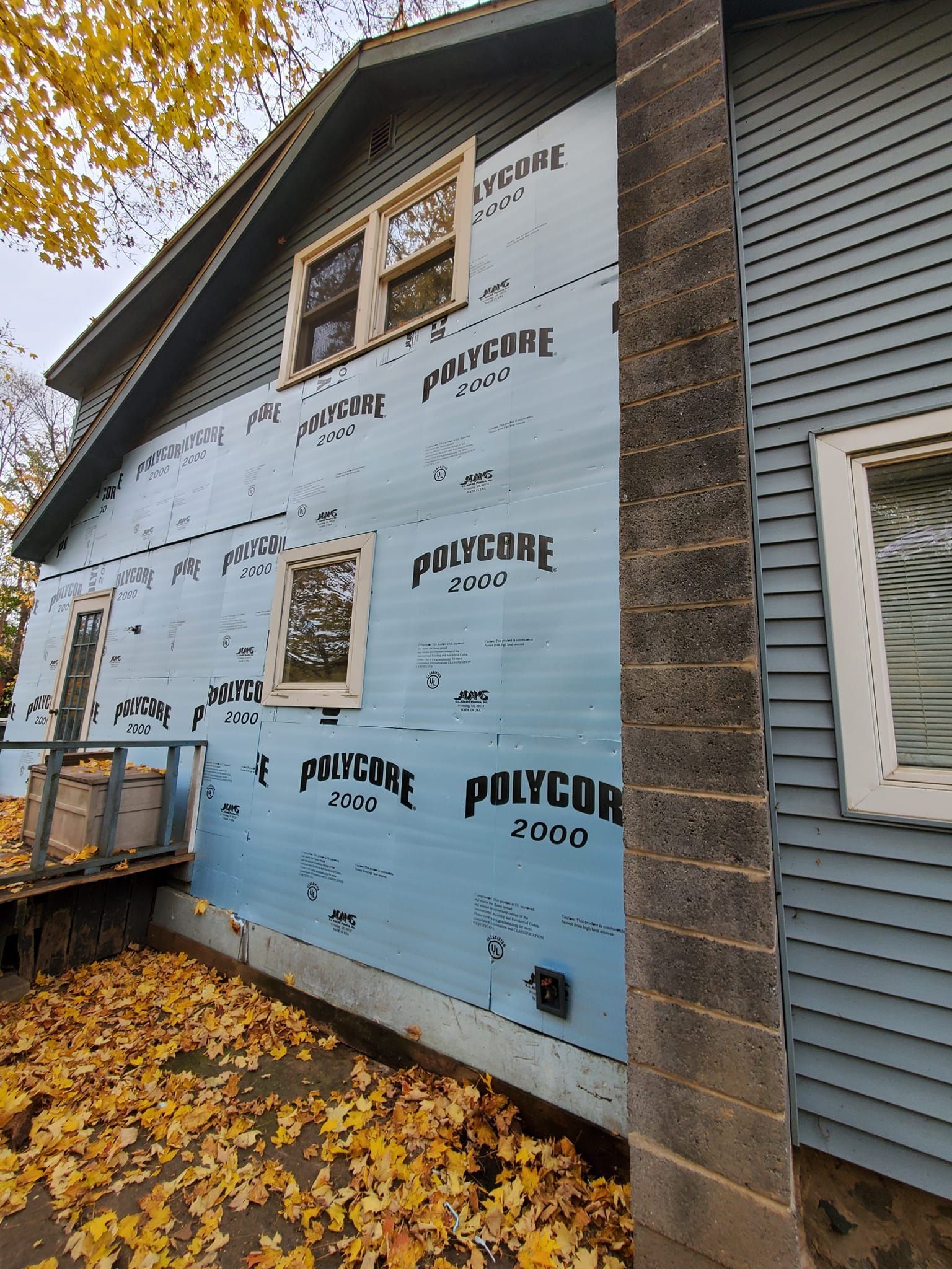 House exterior under renovation with blue siding wrap, windows, and autumn leaves on the ground