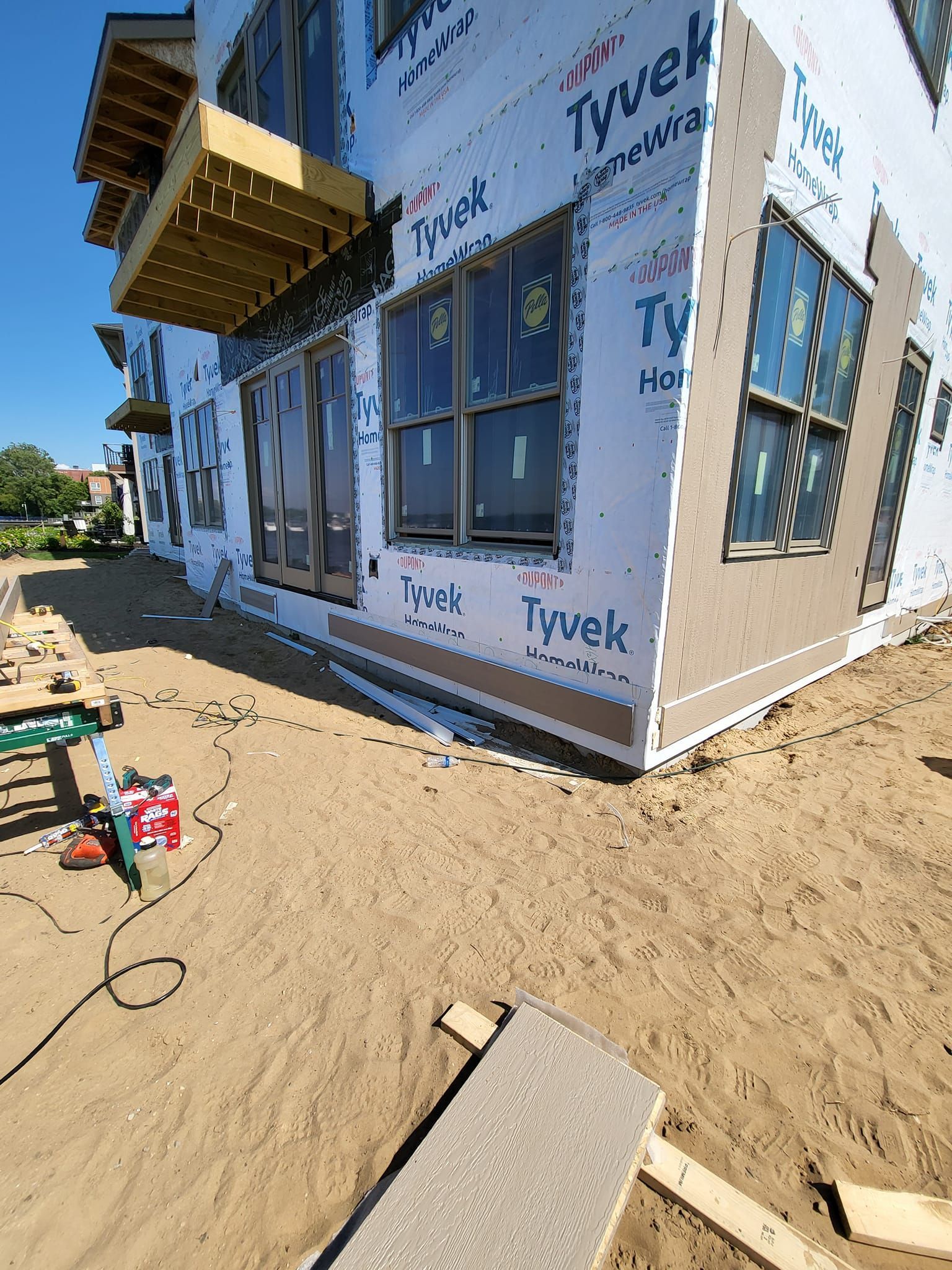 Partly built house wrapped in Tyvek under construction on a sandy lot, with exposed windows and a small porch.