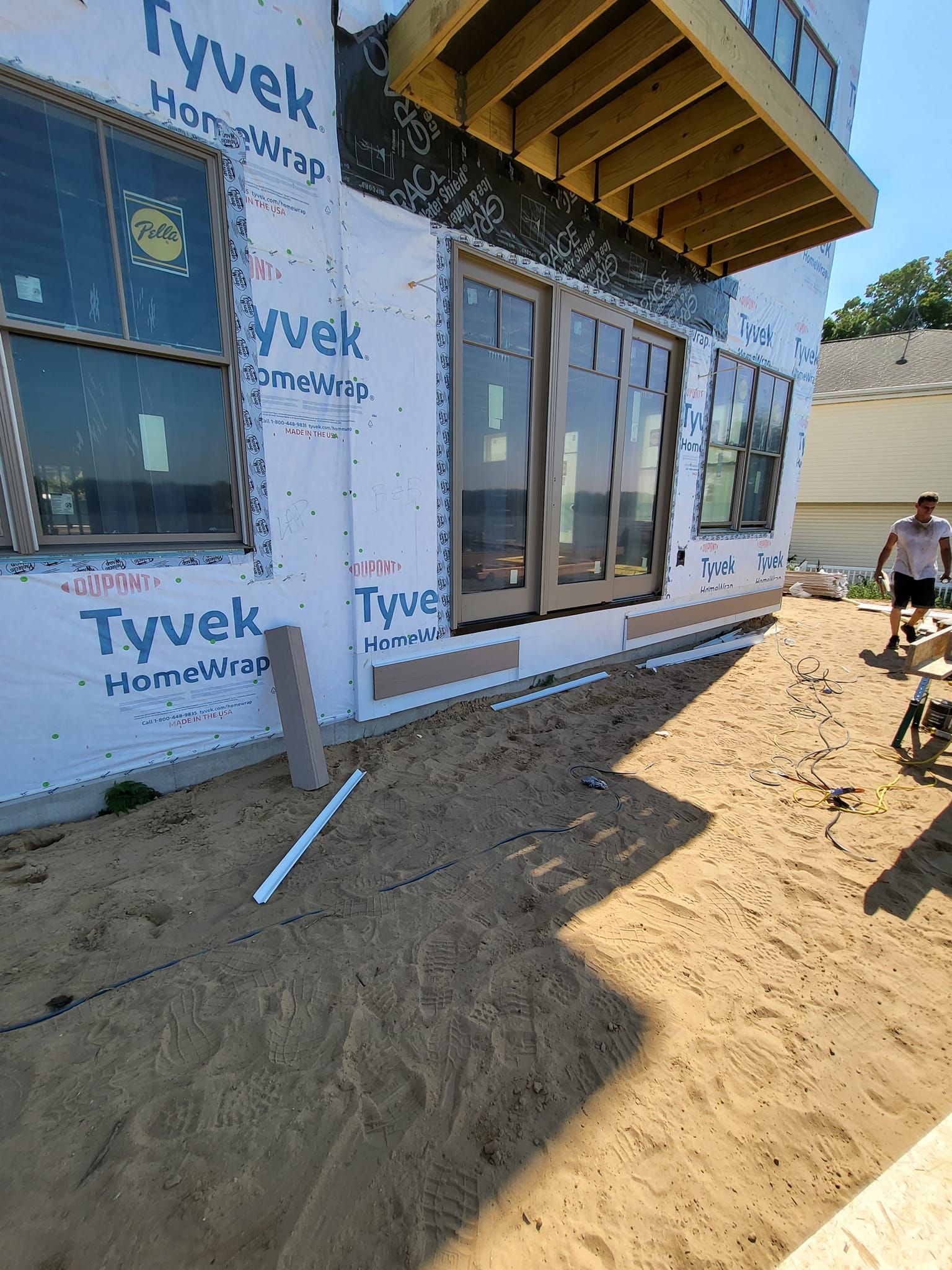 Partially built house wrapped in Tyvek, with windows, sandy yard, and a worker near the side.