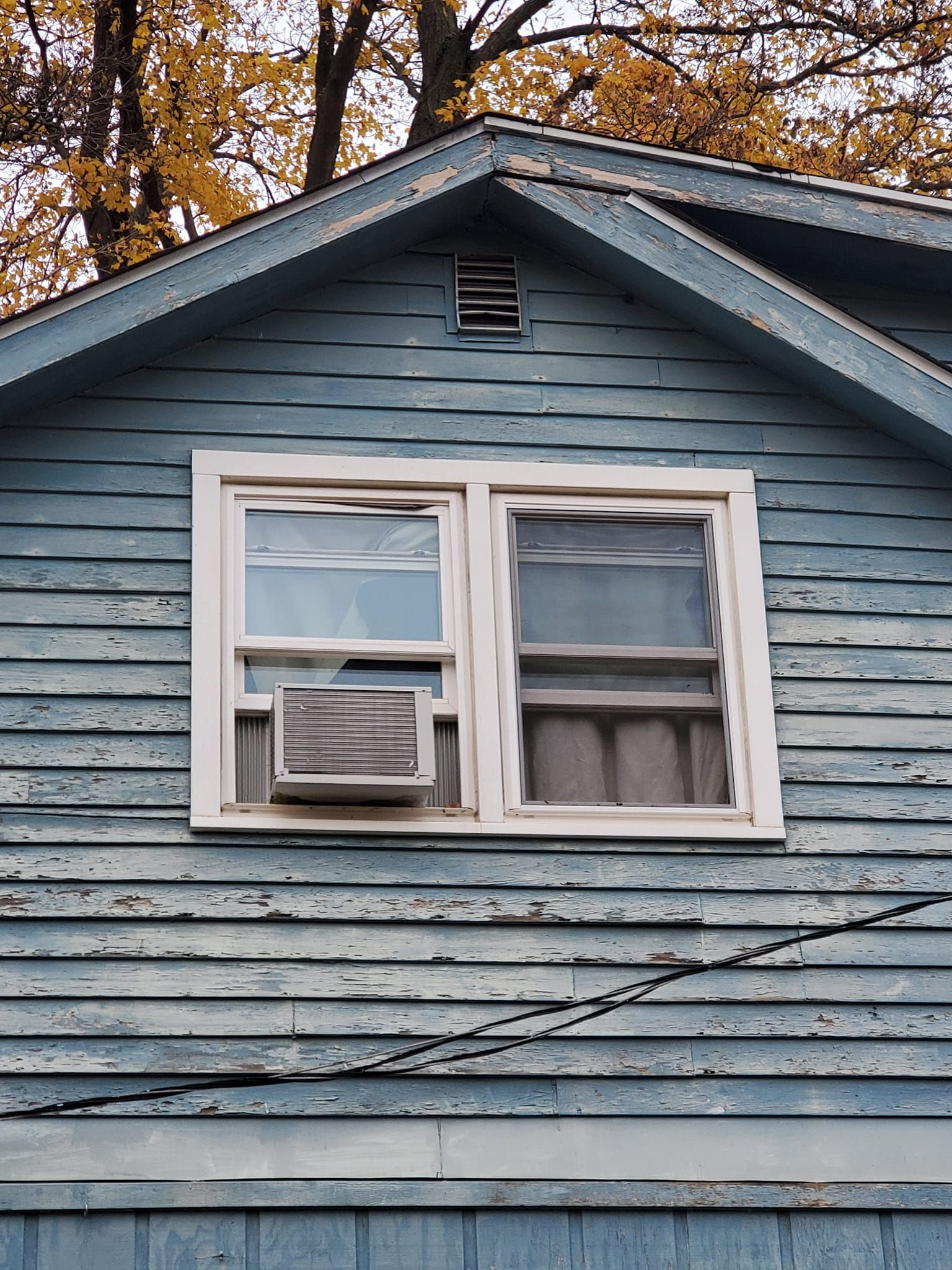 Blue house exterior with a white-framed window air conditioner under a peaked roof in autumn.