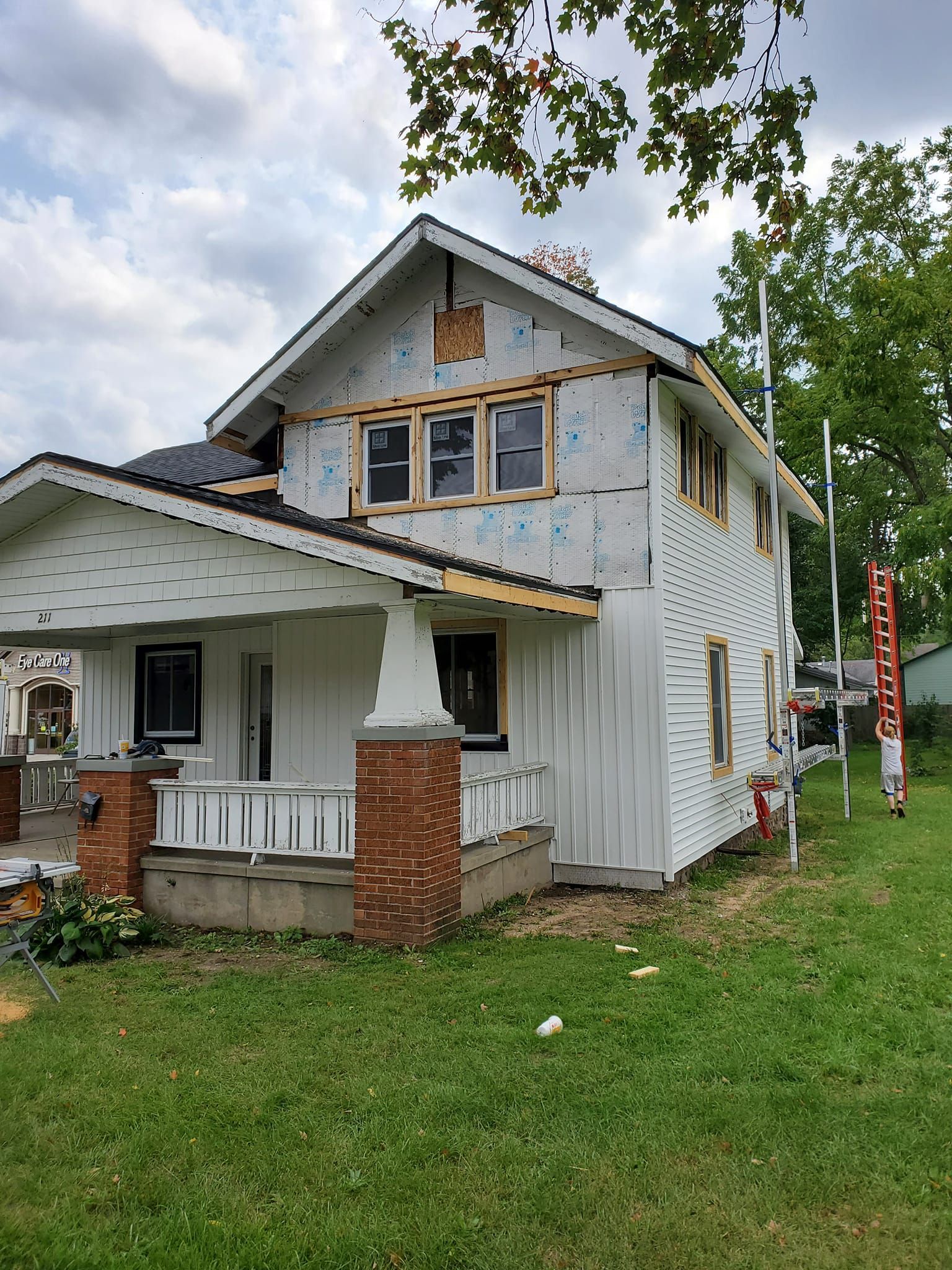 Partially renovated white house with porch, exposed siding, and ladder on a grassy lot