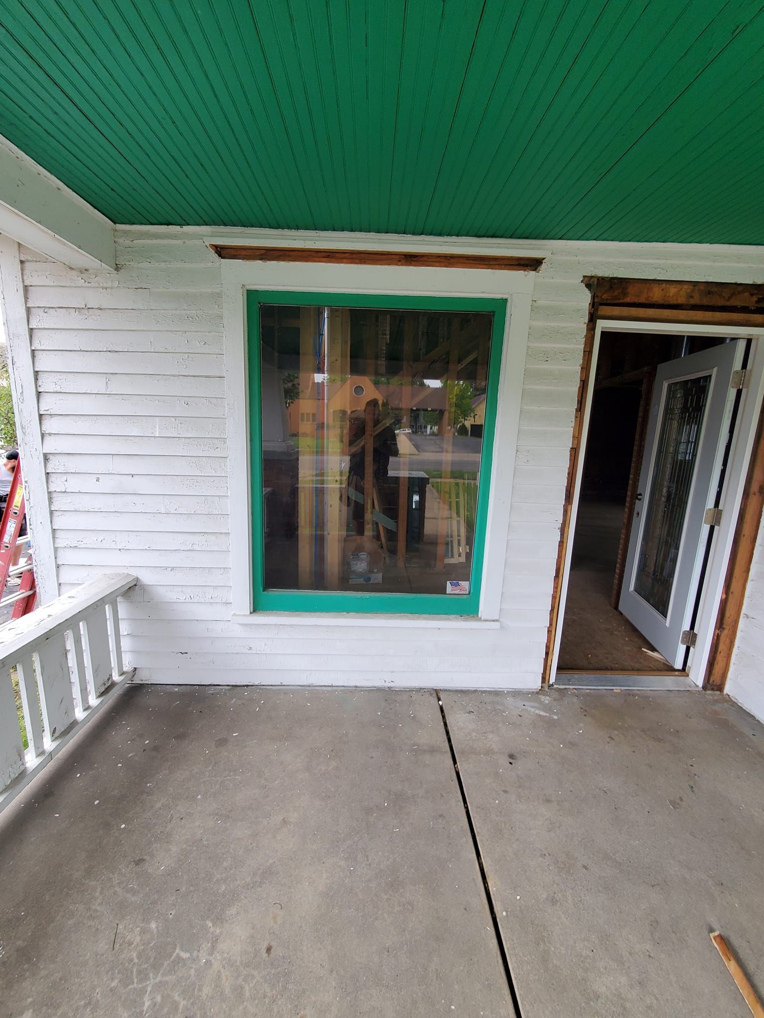 Front porch with white siding, green-trimmed window, open doorway, and concrete floor under a green ceiling.