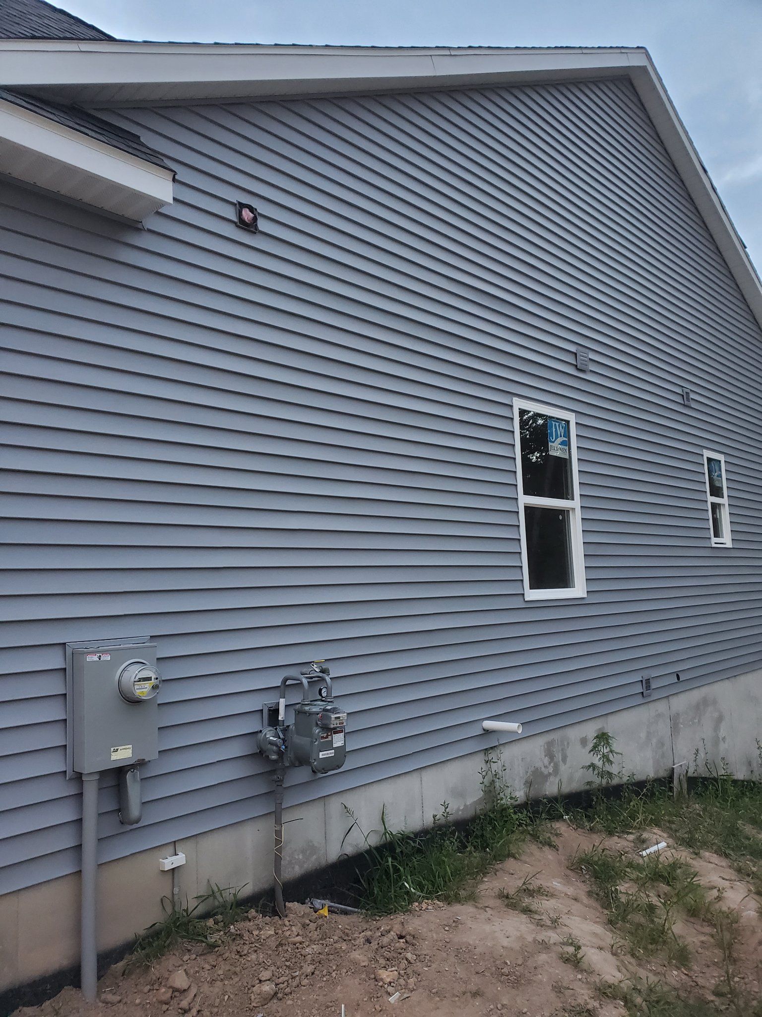 Gray-sided house exterior with two windows, utility boxes, and a dirt strip along the foundation.