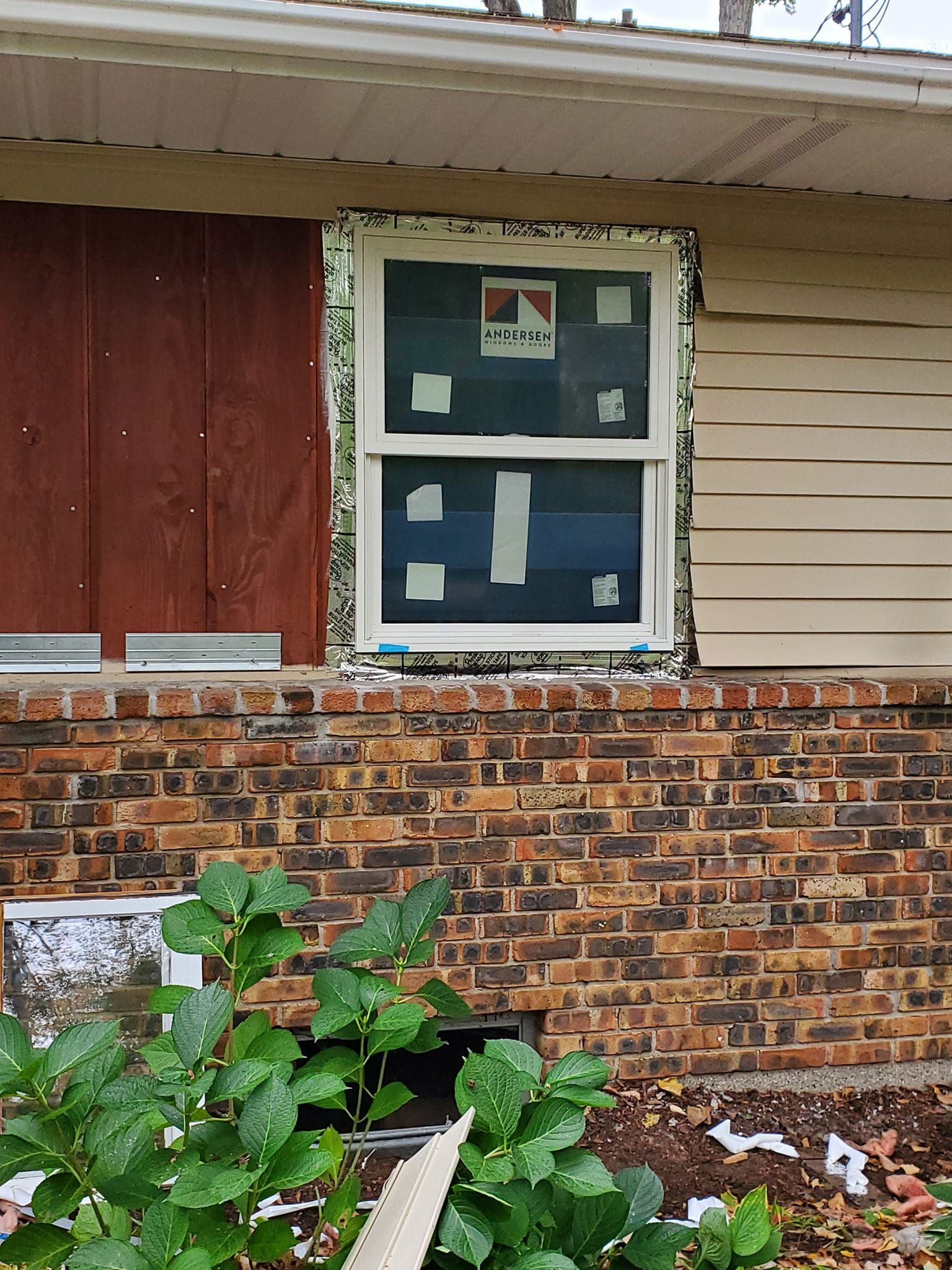 Boarded-up window on a brick house with beige siding and overgrown plants below.