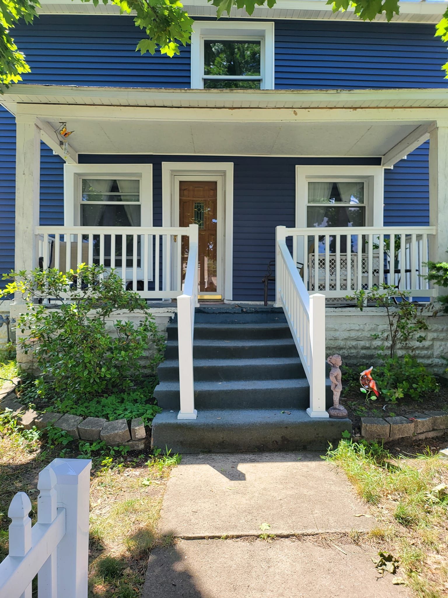 Blue house with white trim, front porch, central steps, and small garden in sunlight