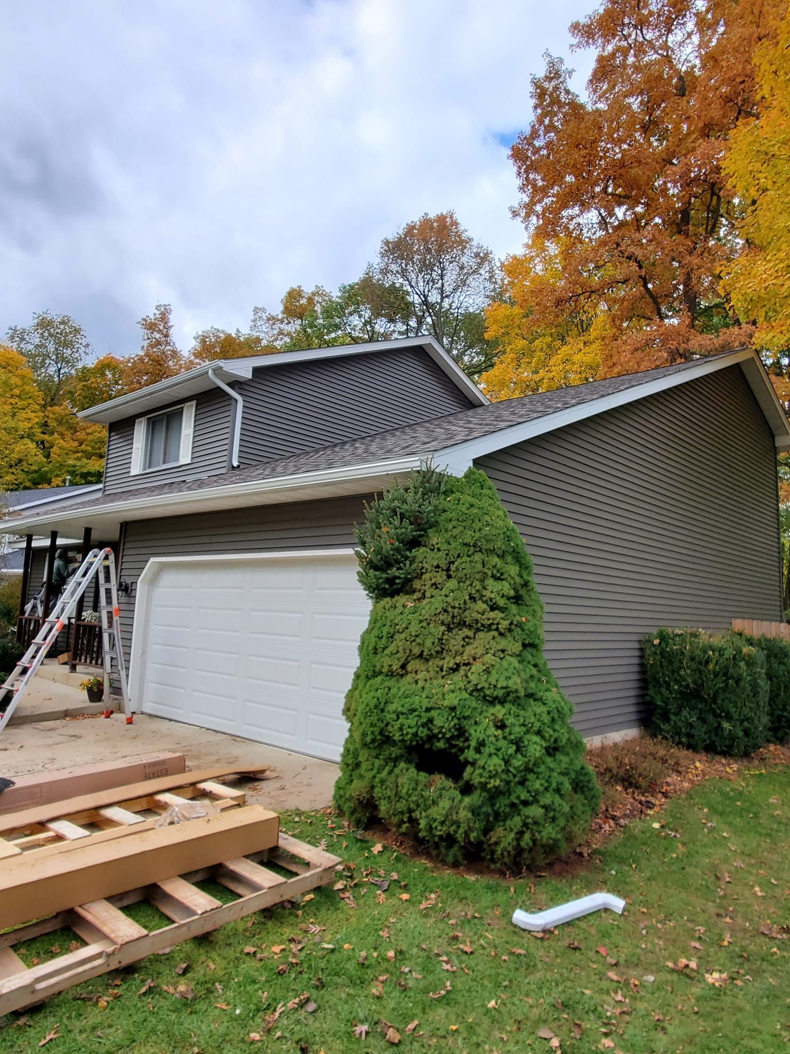 House with damaged siding and garage roof under repair, with ladders and autumn trees nearby