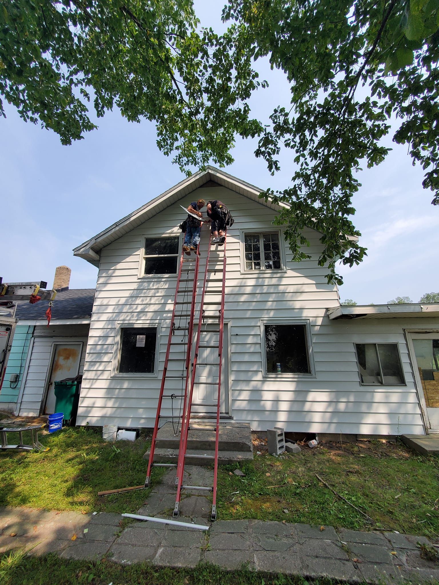 Workers on a tall ladder repairing the front of a white two-story house under tree branches.
