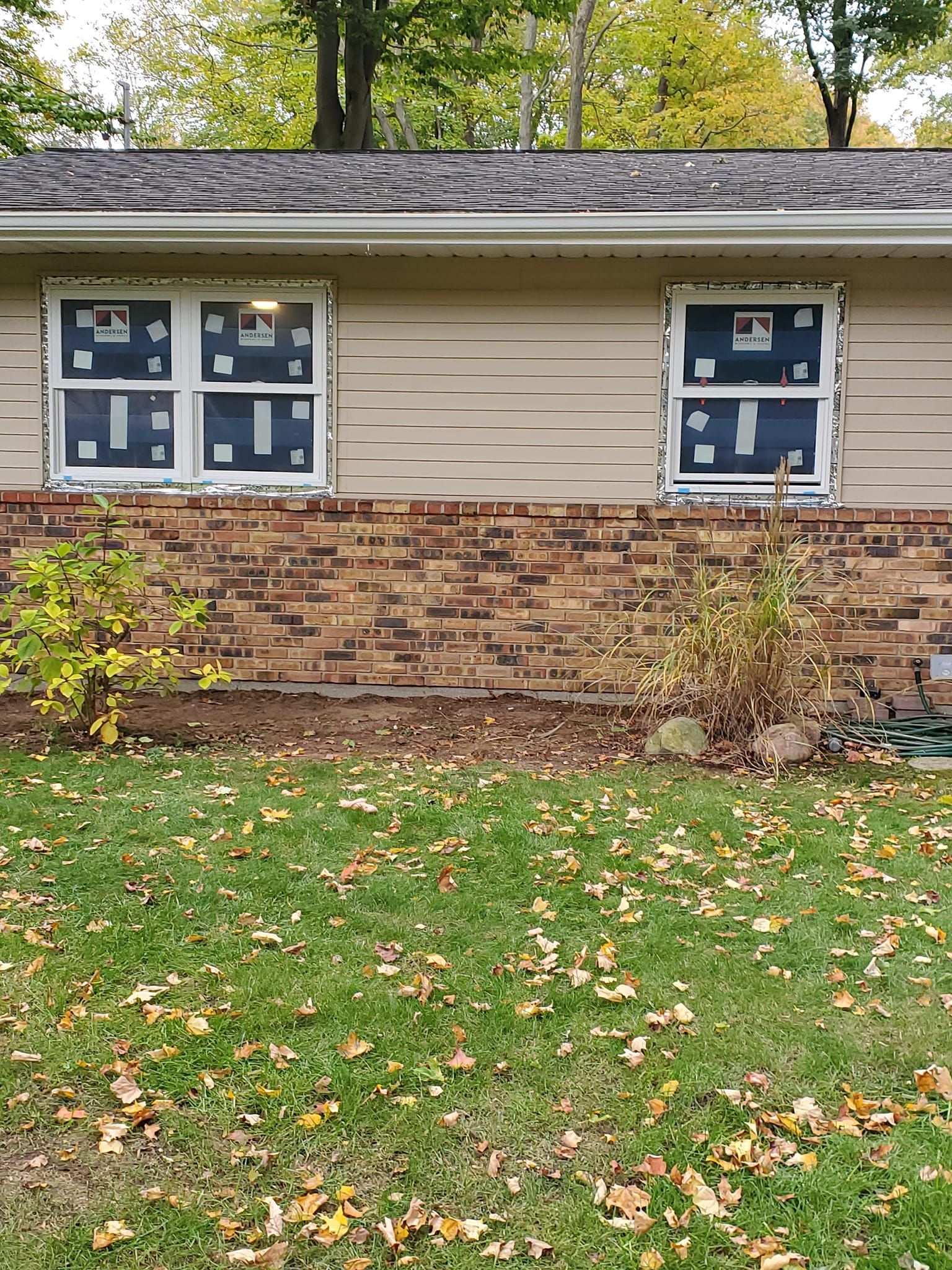 House exterior with tan siding, brick lower wall, and two windows above a grassy lawn in autumn