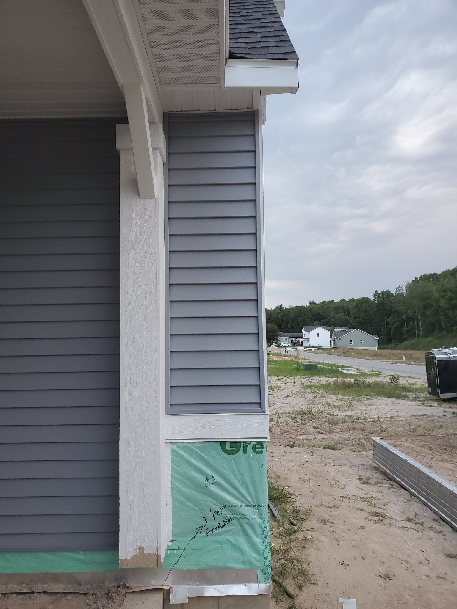 Exterior of a gray house under construction with white trim and green sheathing near a gravel lot