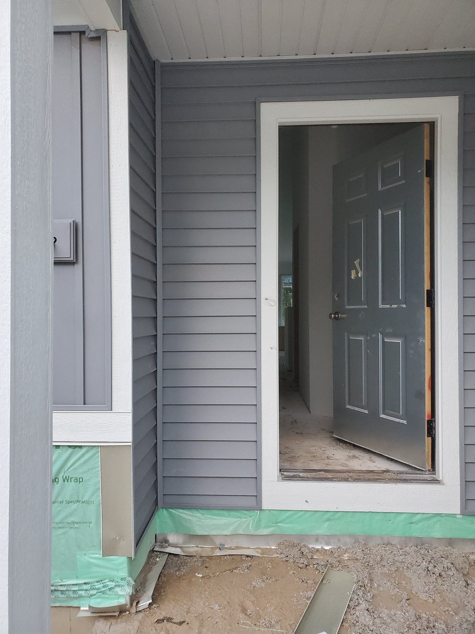 Front porch entry with gray siding, green trim, and an open dark front door over bare ground.