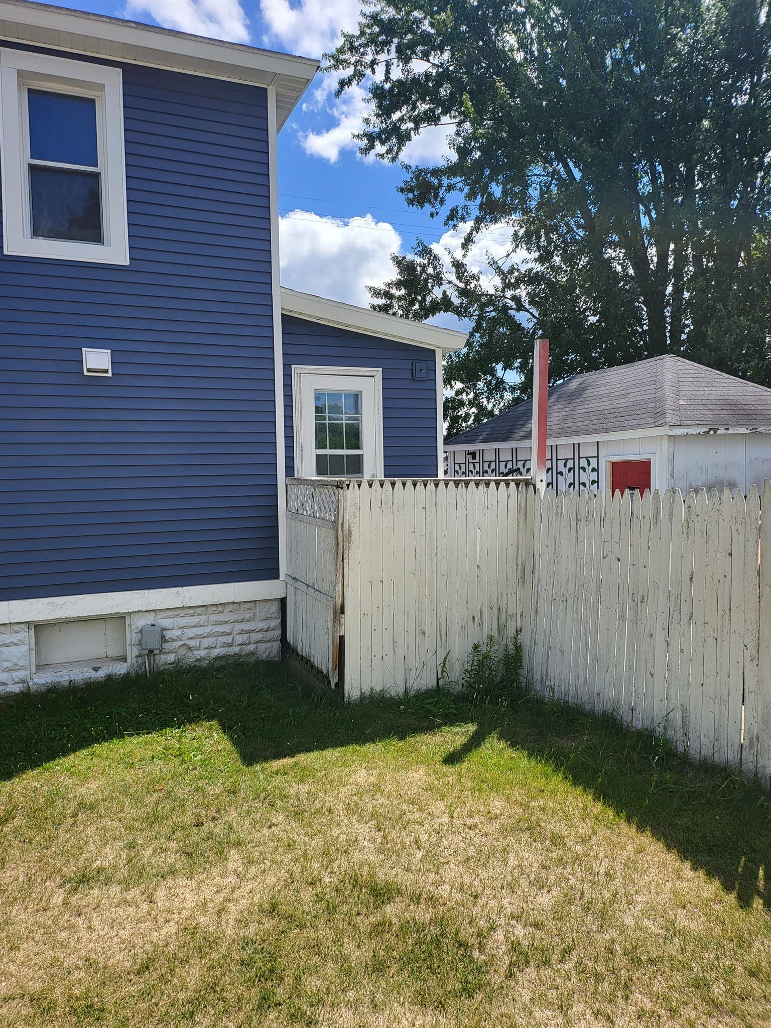 Backyard with blue house, white fence, grass, and a red shed under a partly cloudy sky