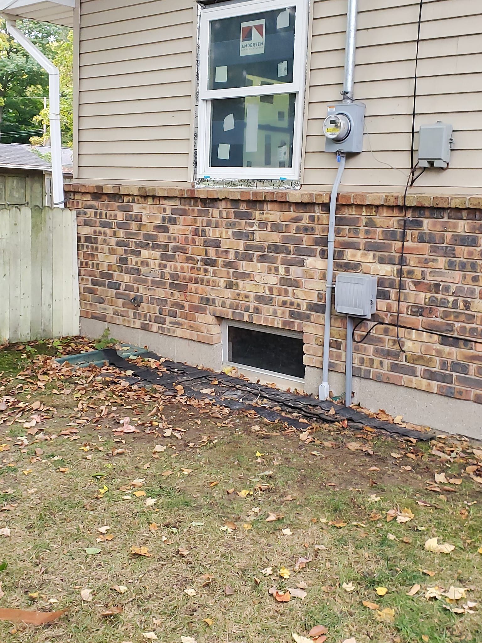 House exterior with tan siding, brick veneer, utility meters, and a basement vent near a grassy yard.