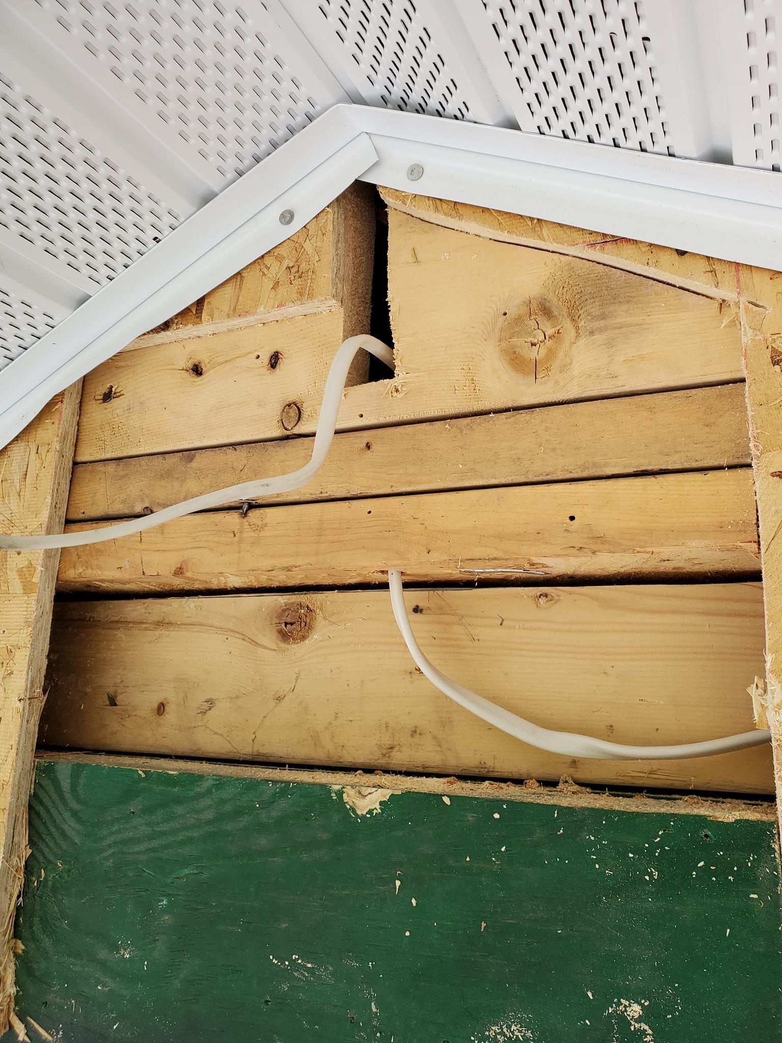 Damaged wooden wall under a white perforated soffit with loose white cable and green base trim