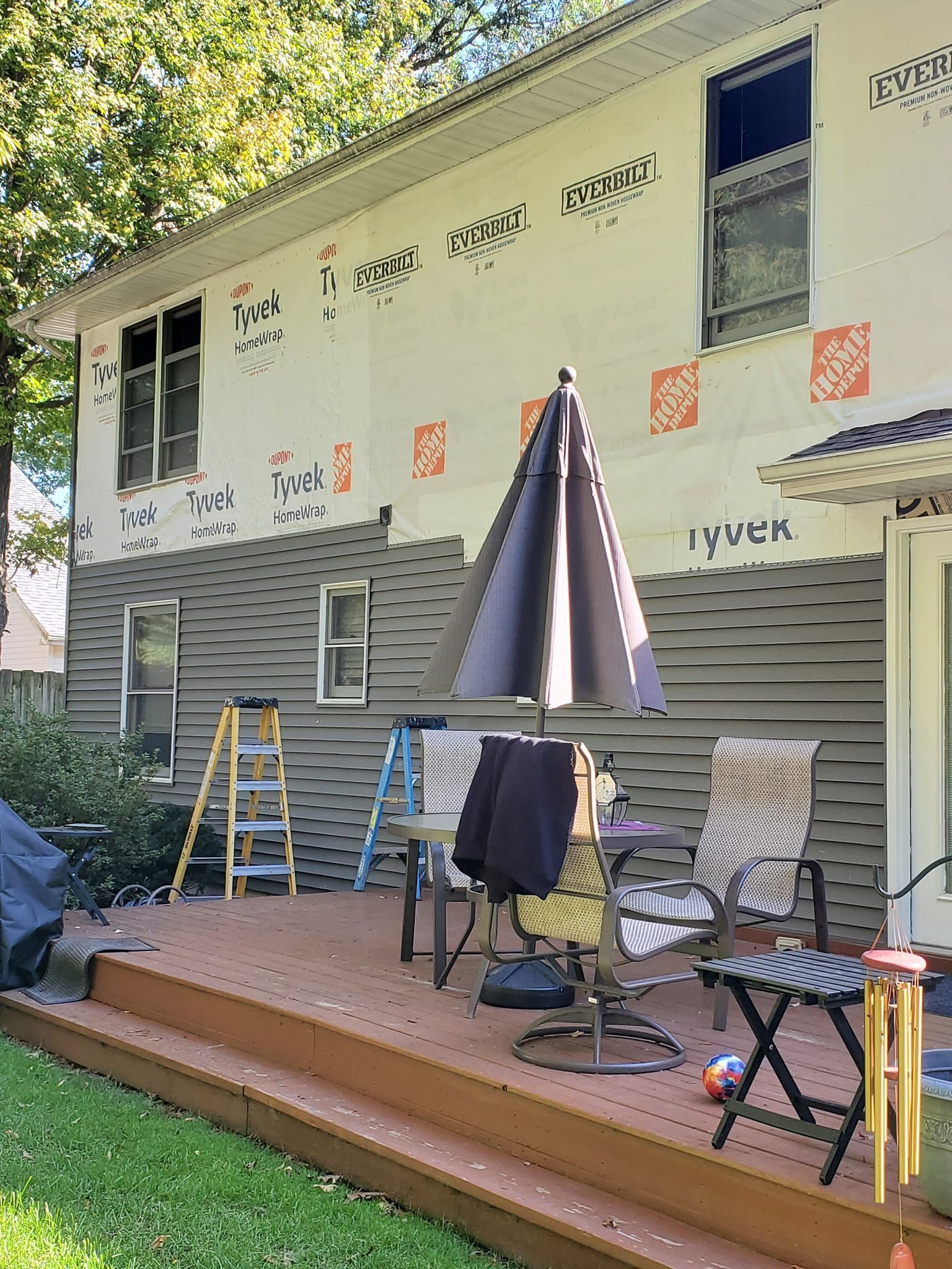Outdoor patio with folding chairs, a table, umbrellas, ladders, and construction materials beside a house