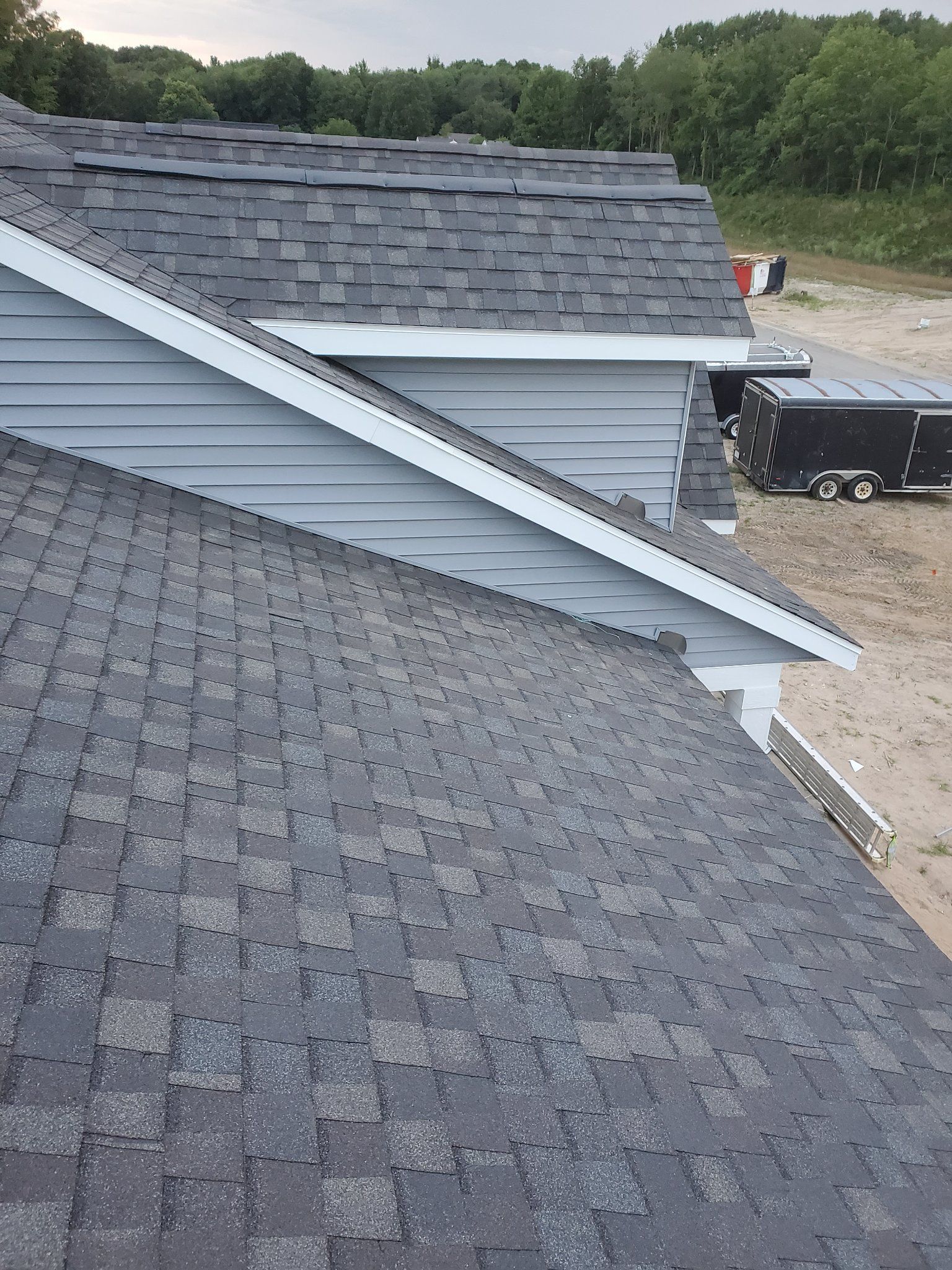 Roof shingles on a house under construction, with white siding and a gravel worksite in the background.