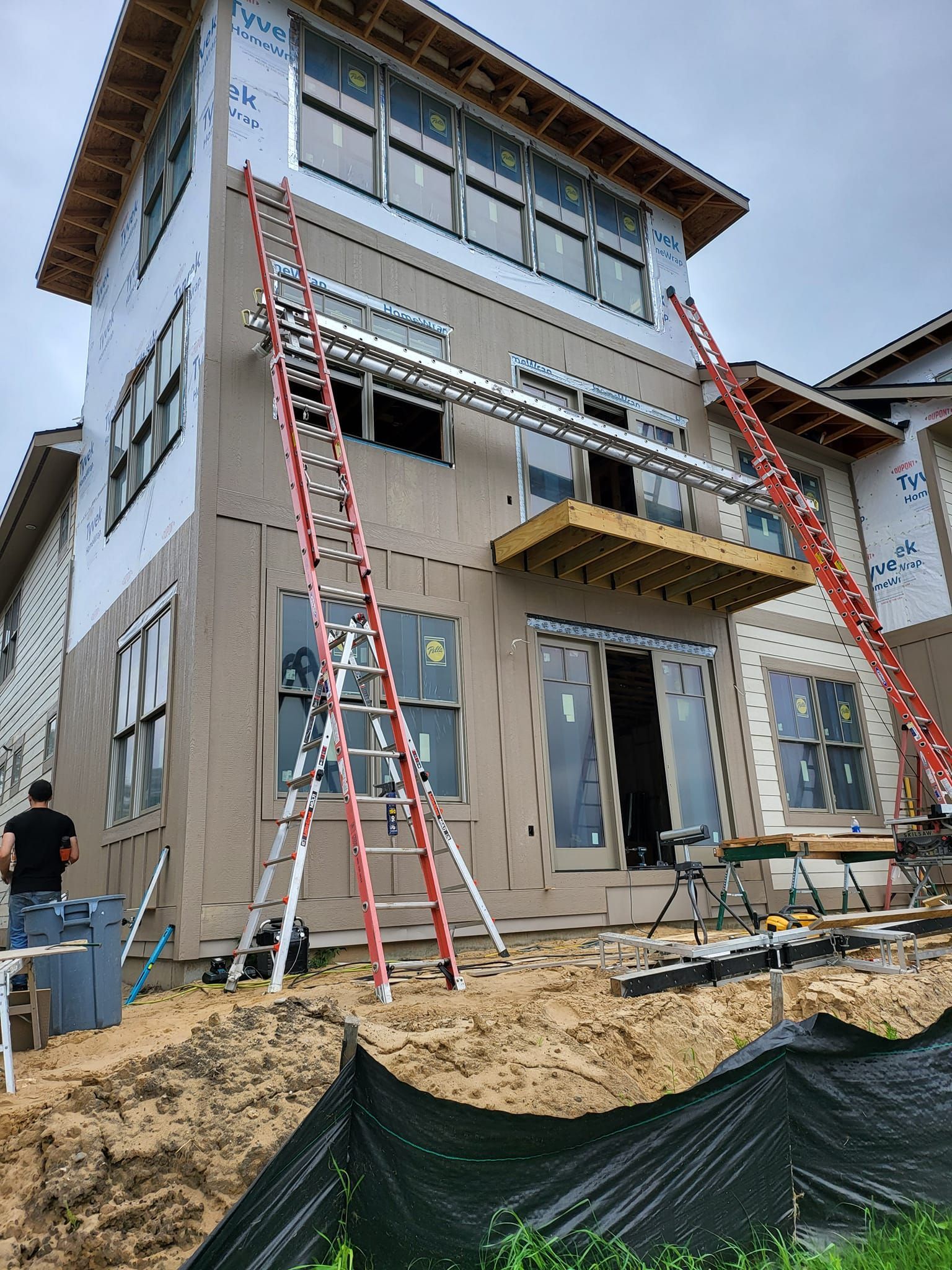 New house under construction with ladders, scaffolding, and workers on a dirt lot
