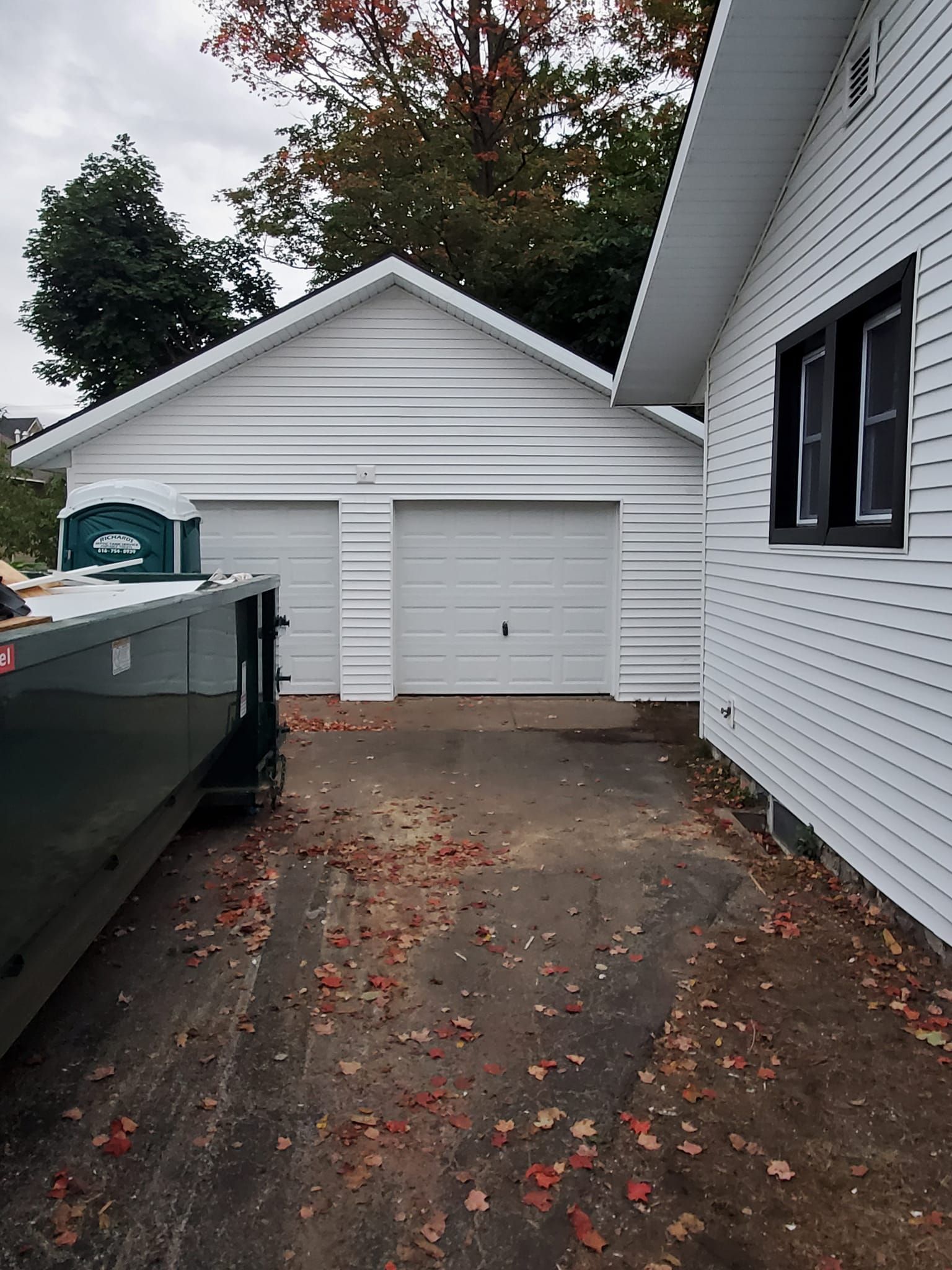 Driveway beside a white house leading to a detached garage with double doors and a dumpster on the left.