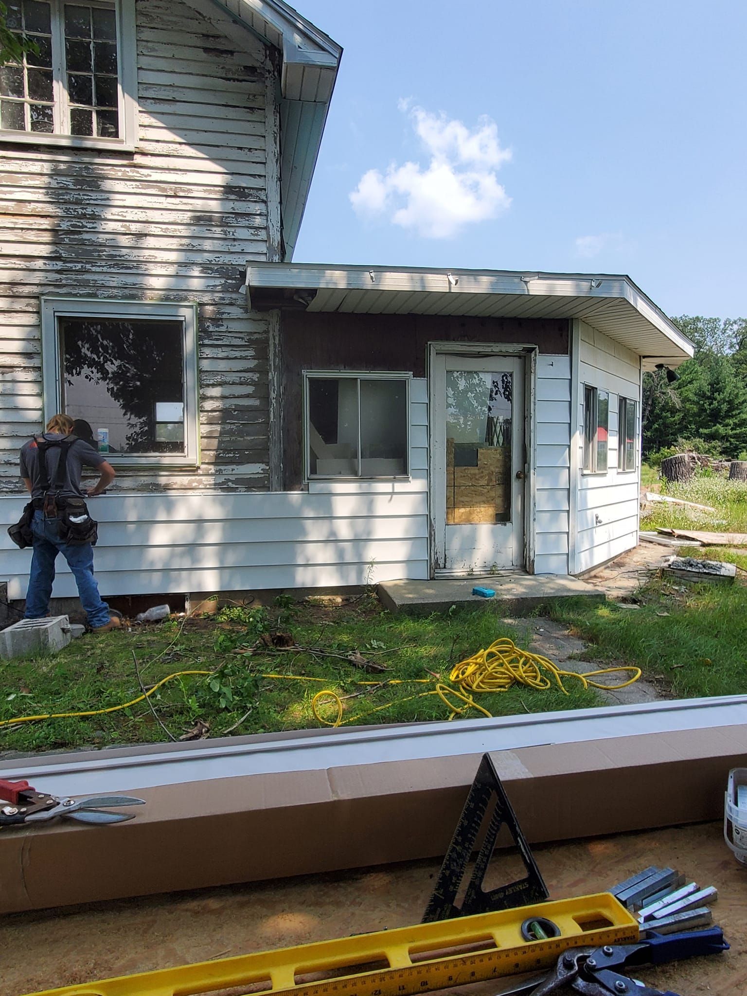 Worker standing by a white house with stone siding; tools in foreground and a sunny sky.
